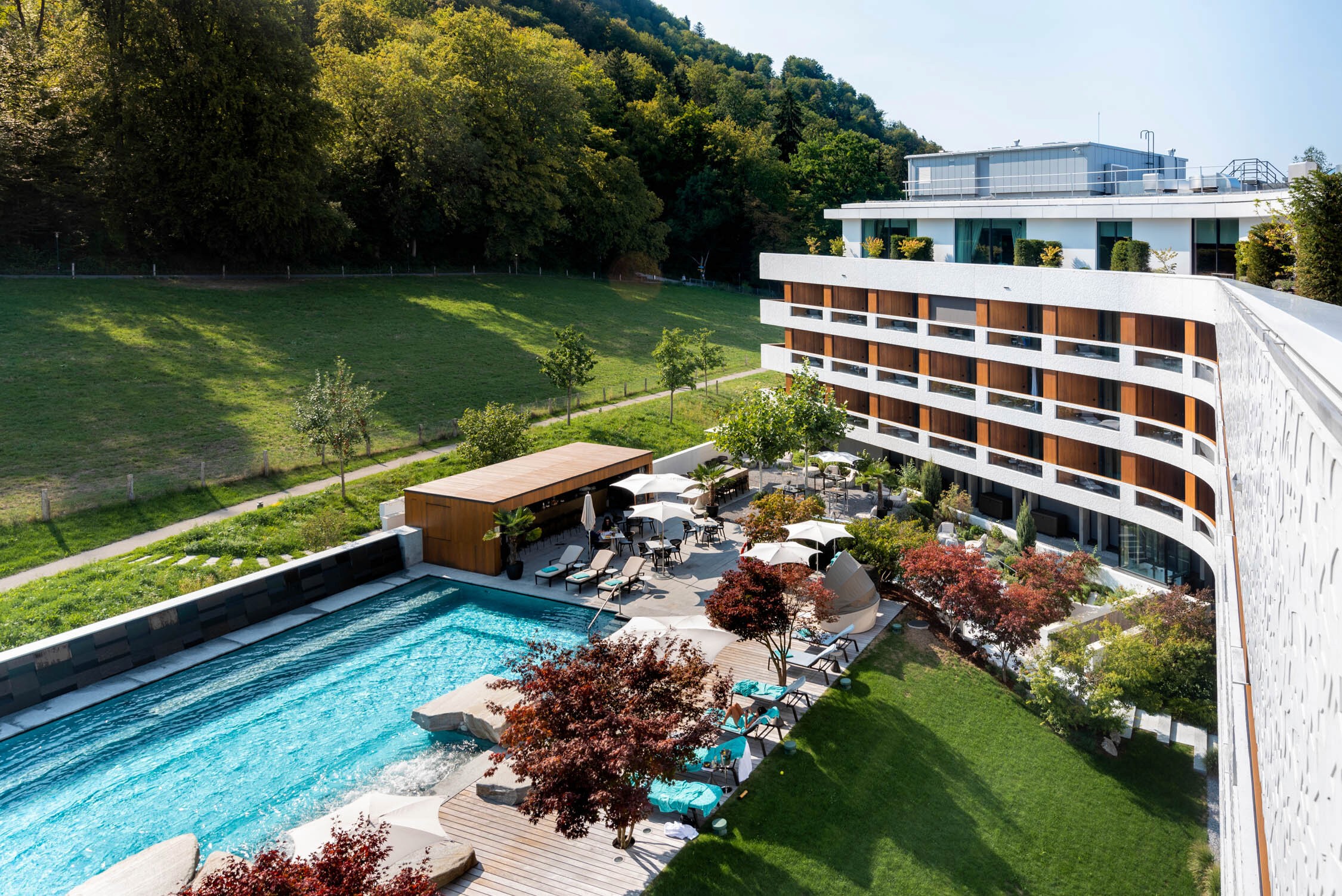 A modern hotel with a long balcony overlooks a swimming pool and lounge area set against a lush green hillside.
