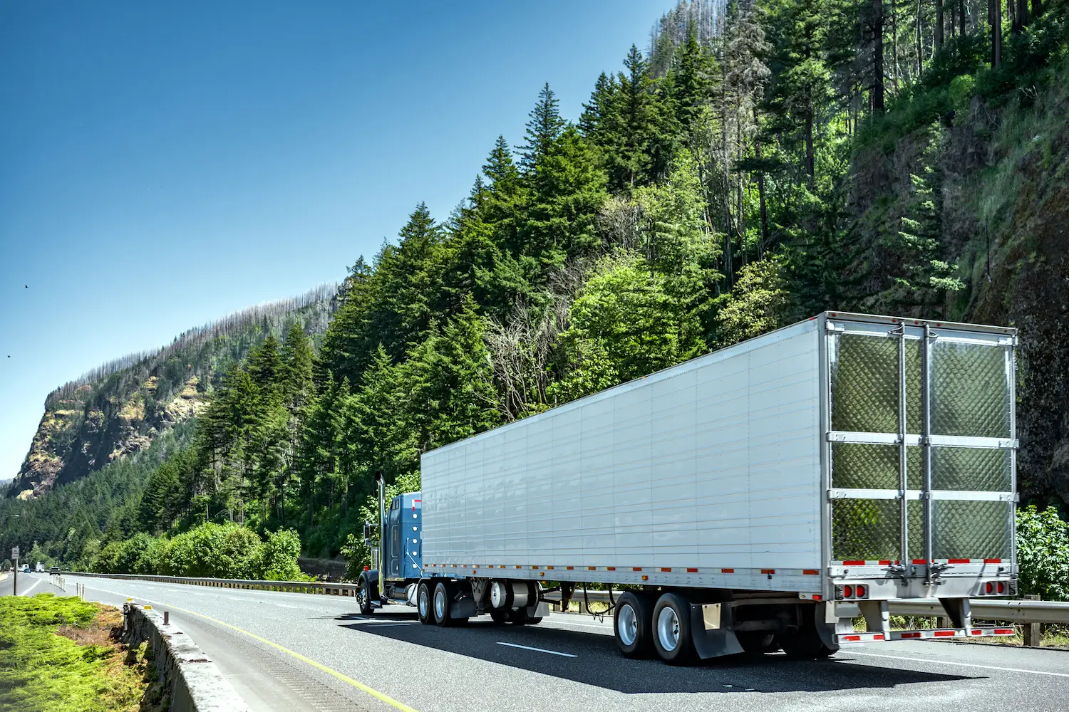 Blue semi-truck with a white trailer driving through a scenic mountain highway surrounded by pine trees, representing successful trucking operations insured by GIA Group, LLC.