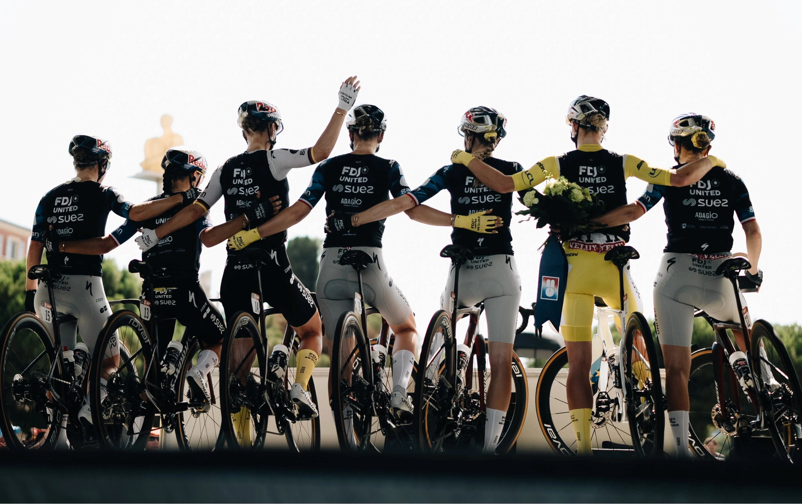 FDJ United-SUEZ riders lined up arm in arm on their bikes after the Tour de France Femmes, the race leader in the yellow jersey holding a winner's bouquet.