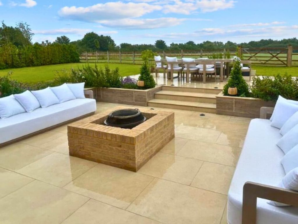 A cozy outdoor seating area with white sofas around a square fire pit, overlooking a green landscape.