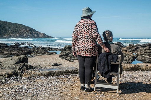 An older female stood next to a man in a wheelchair, with her arm on the back of the chair. They are at the beach and looking out to a cliff and the ocean.