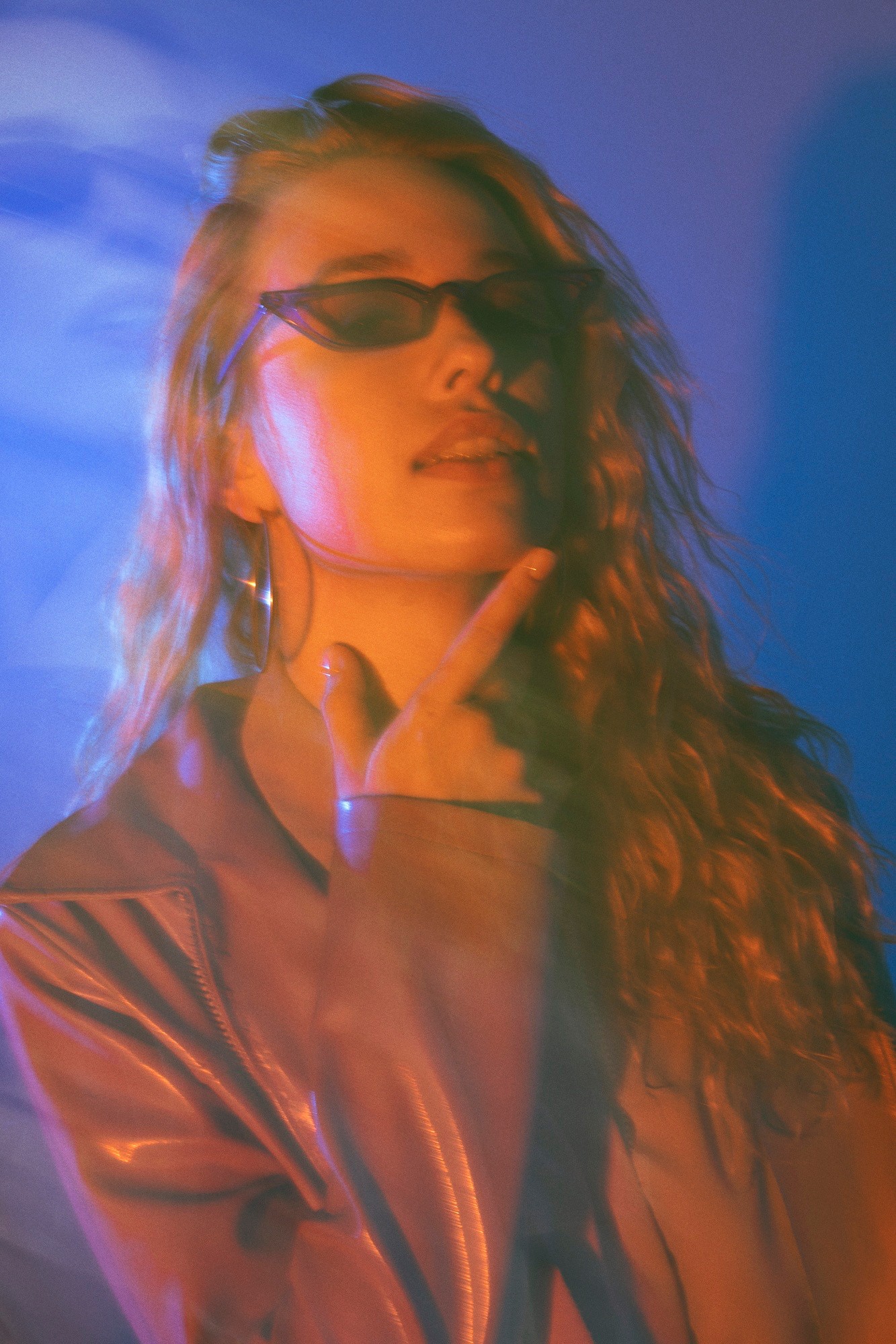 Young woman with blonde-red hair wearing sunglasses and a red leather jacket, standing against a blue background.