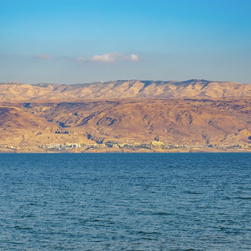 A calm blue sea with a background of arid, mountainous terrain under a clear blue sky.