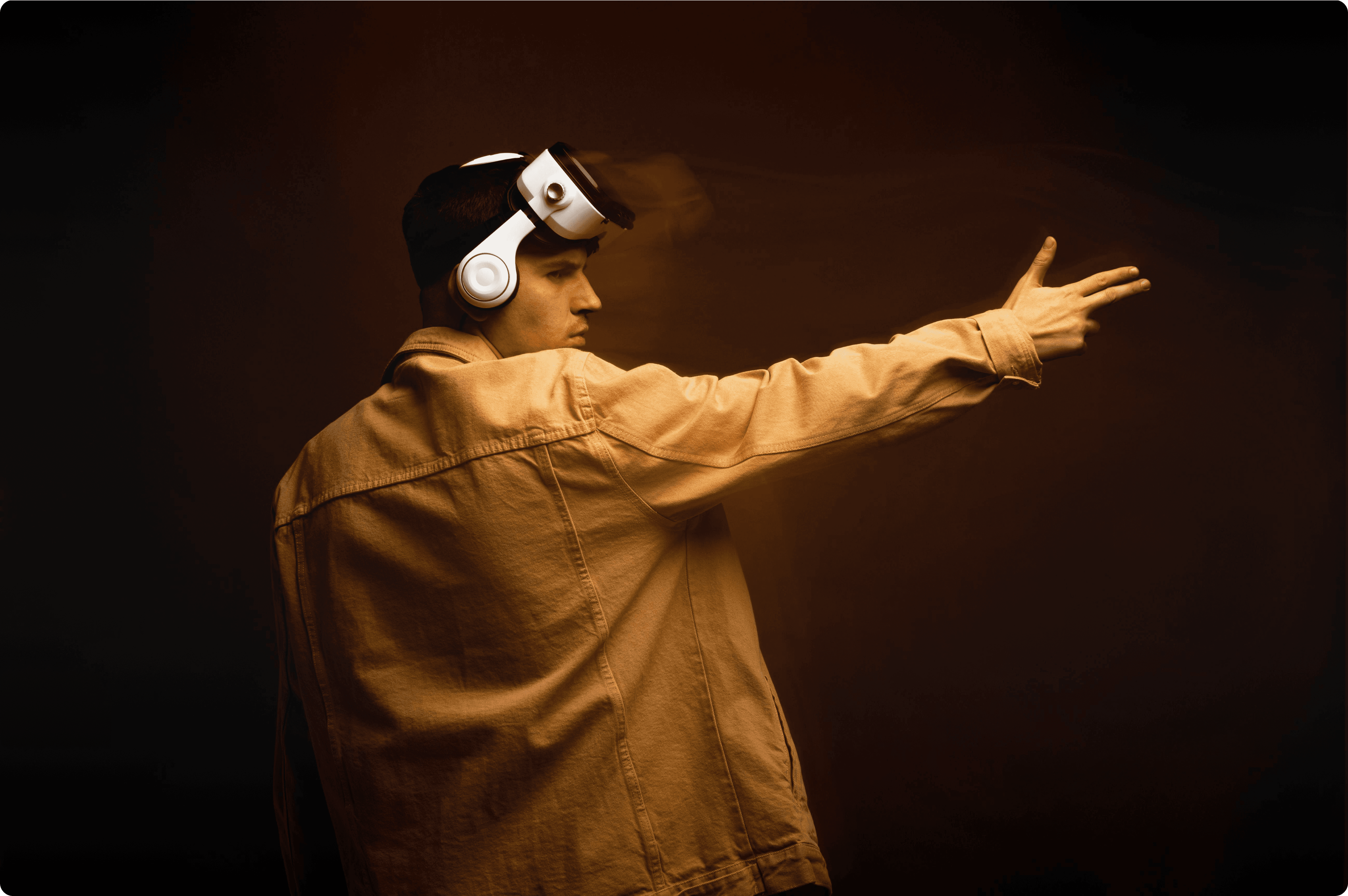A cinematic side profile of a man wearing a white VR headset and a mustard yellow denim jacket. He is pointing his hand forward in a shooting gesture against a dark, moody amber background with atmospheric smoke and warm studio lighting.