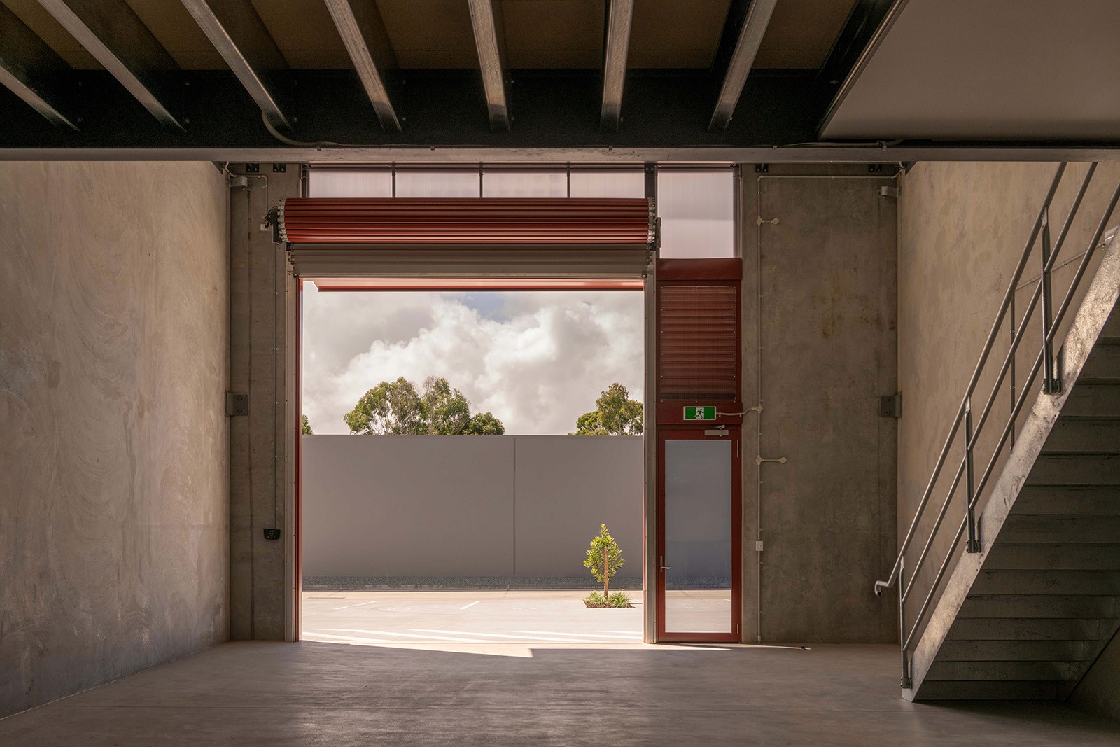 Interior of a RedSheds unit looking out through an open red roller door, revealing raw concrete walls, exposed structure, a steel stair, and a clear connection to the outdoor yard beyond.