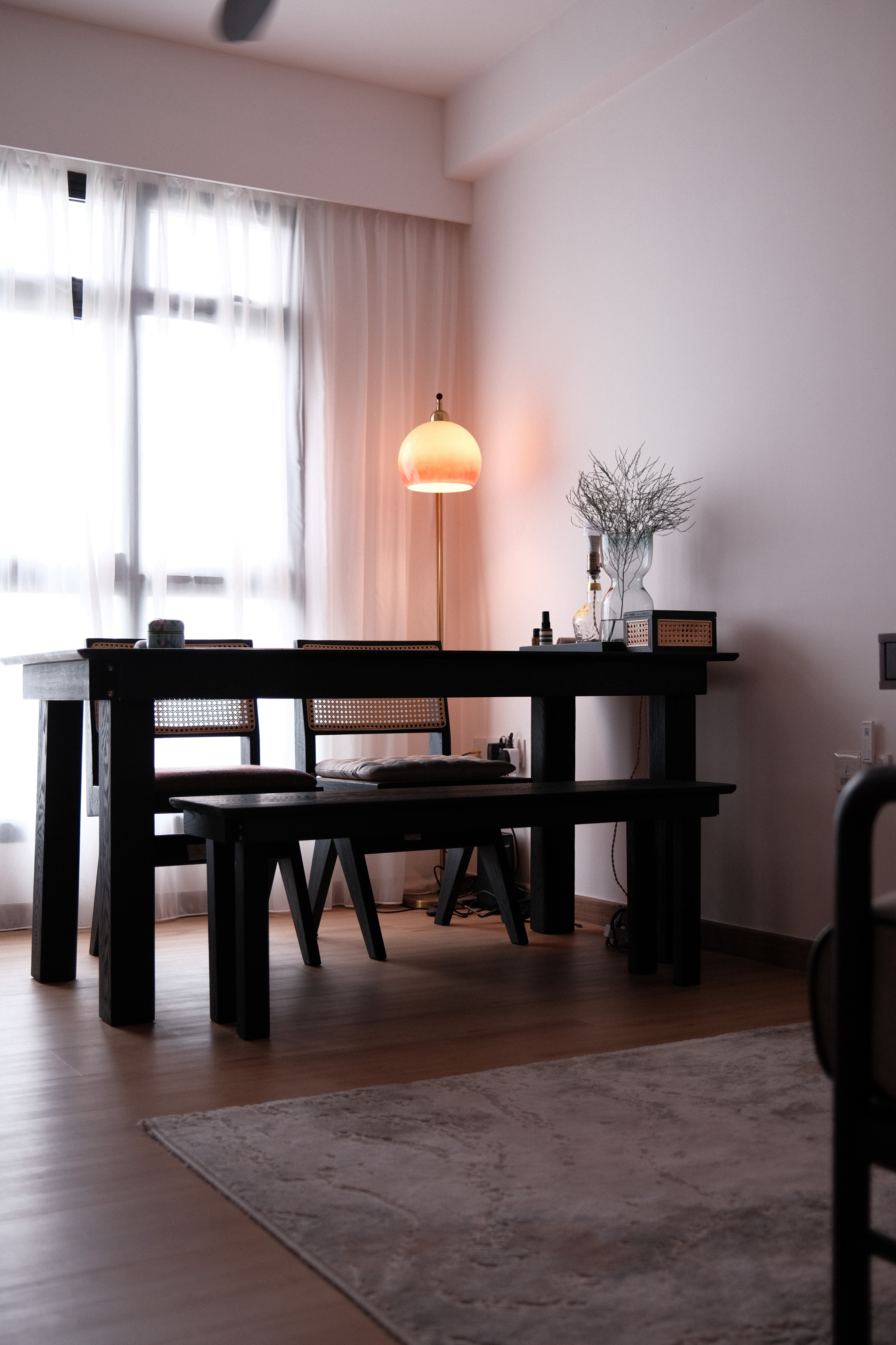 Contemporary dining area with black wood table, woven rattan chairs, glass vase with foliage, and a brass arc floor lamp softly lighting the space.