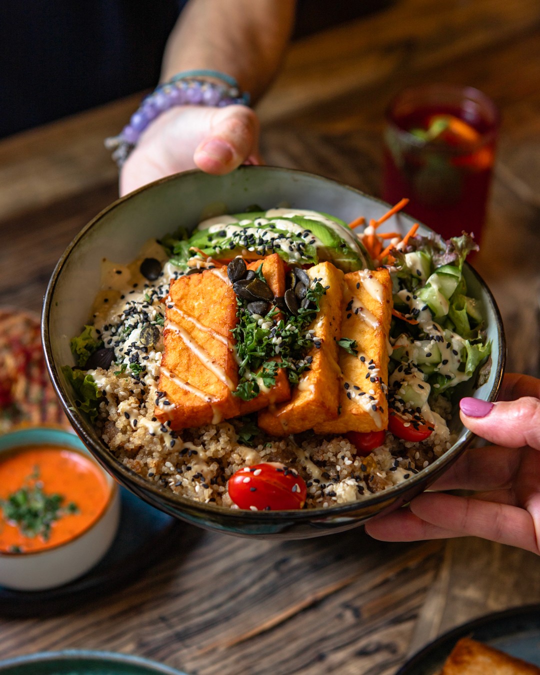 Hände präsentieren eine große Schüssel gefüllt mit bunten Zutaten wie geröstetem Gemüse, Getreide, Kichererbsen und einer leuchtenden Sauce.
