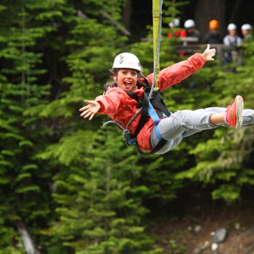 一個穿著紅色夾克和頭盔的人正快樂地在樹林區域進行滑索，背景中有其他戴著頭盔的人。