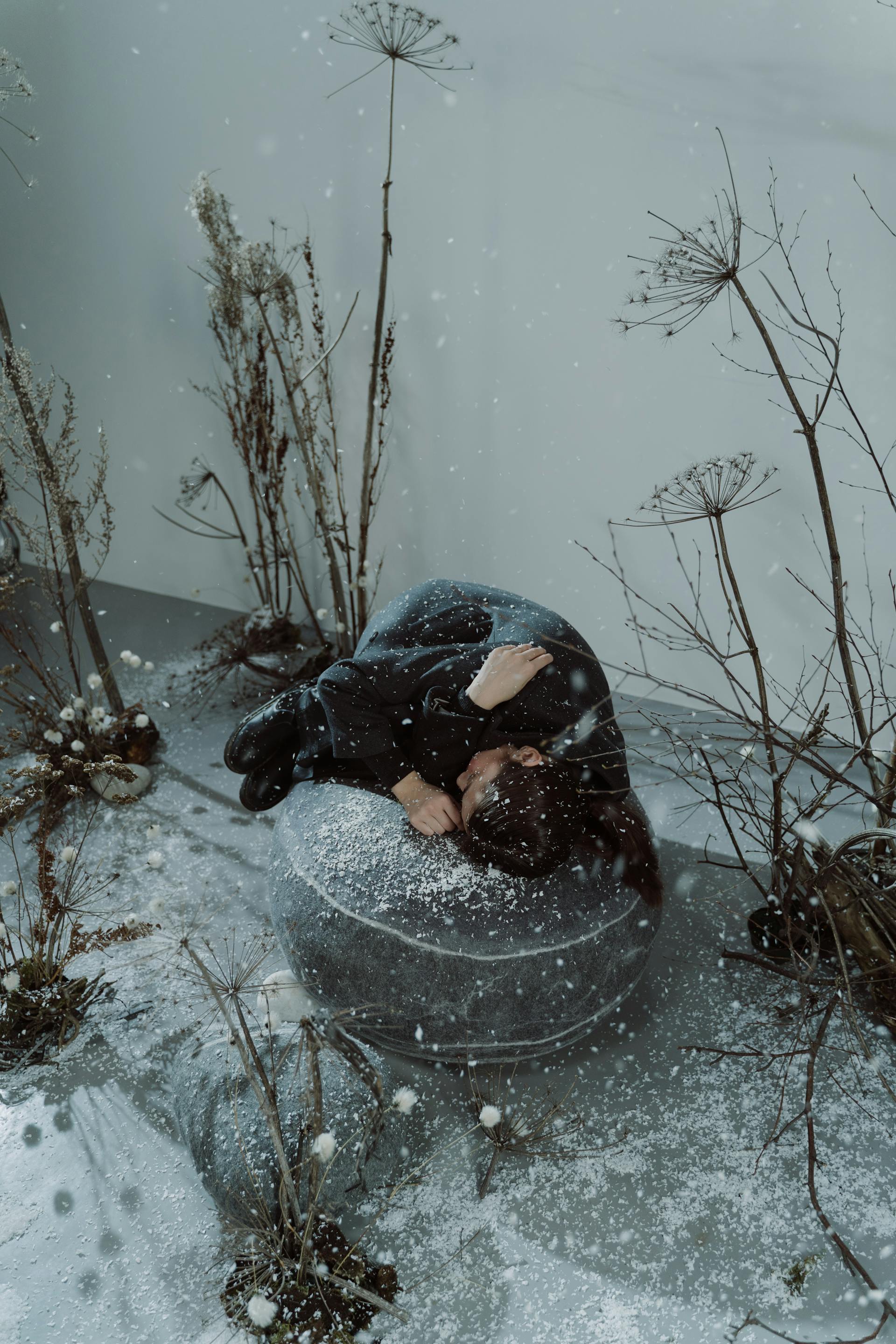 A high-angle shot showing a woman curled up on a large, snow-covered stone or beanbag chair in a minimalist studio setting with falling snow and dried flora.