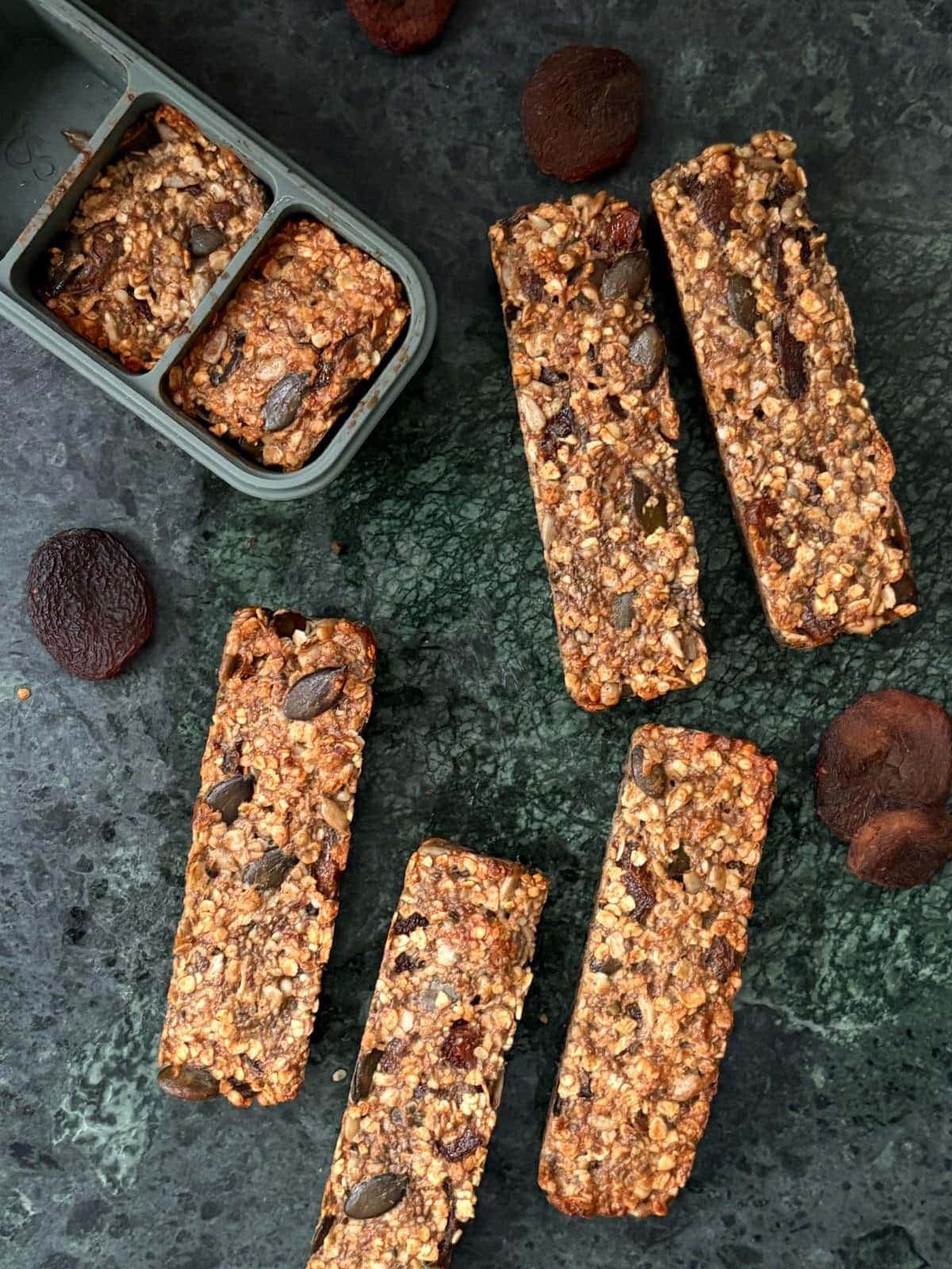 Homemade apricot oat bars with visible pumpkin seeds and dried fruit, arranged on a dark surface with whole apricots nearby.