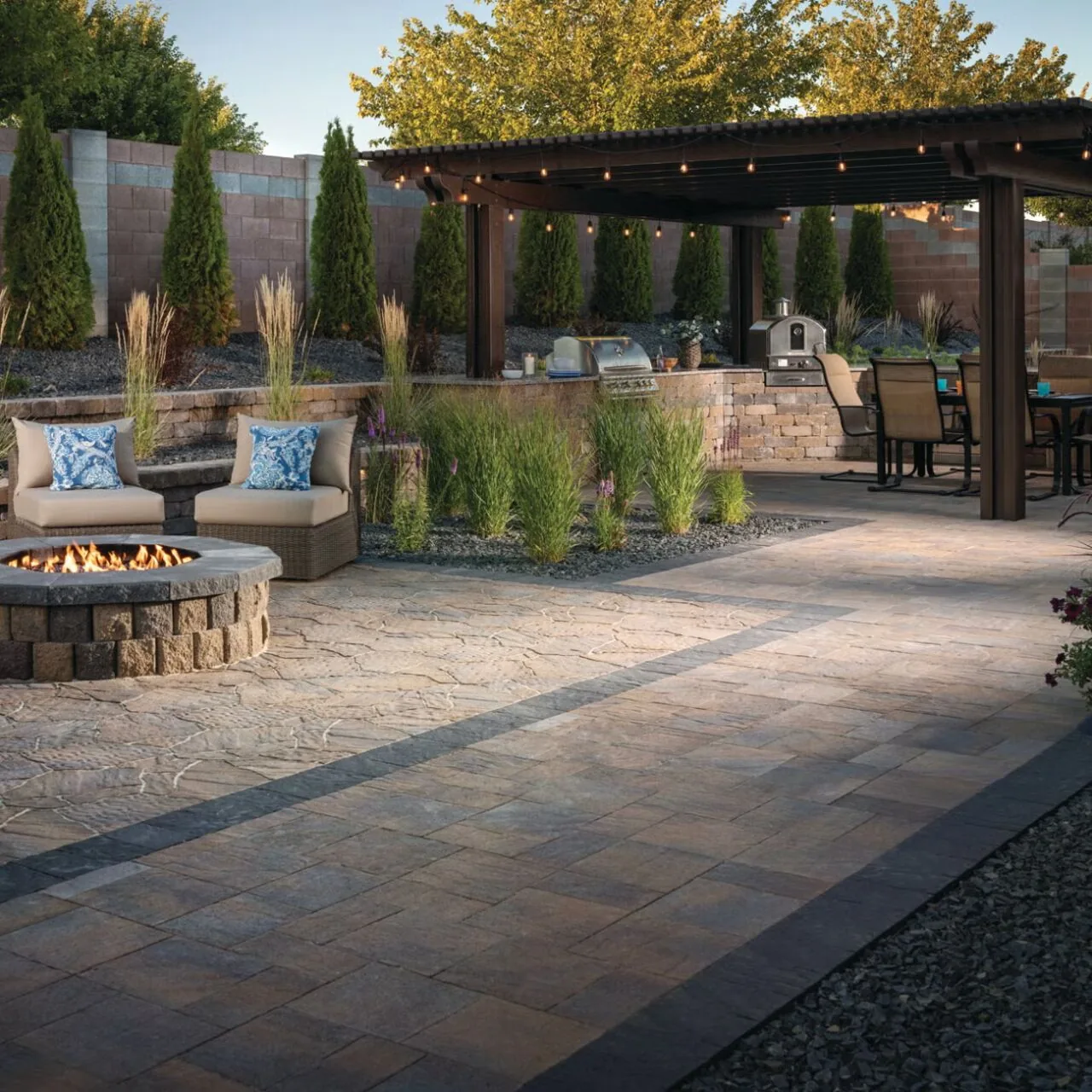 Paved patio with a round fire pit, lounge chairs, planted borders and a pergola over an outdoor dining area.