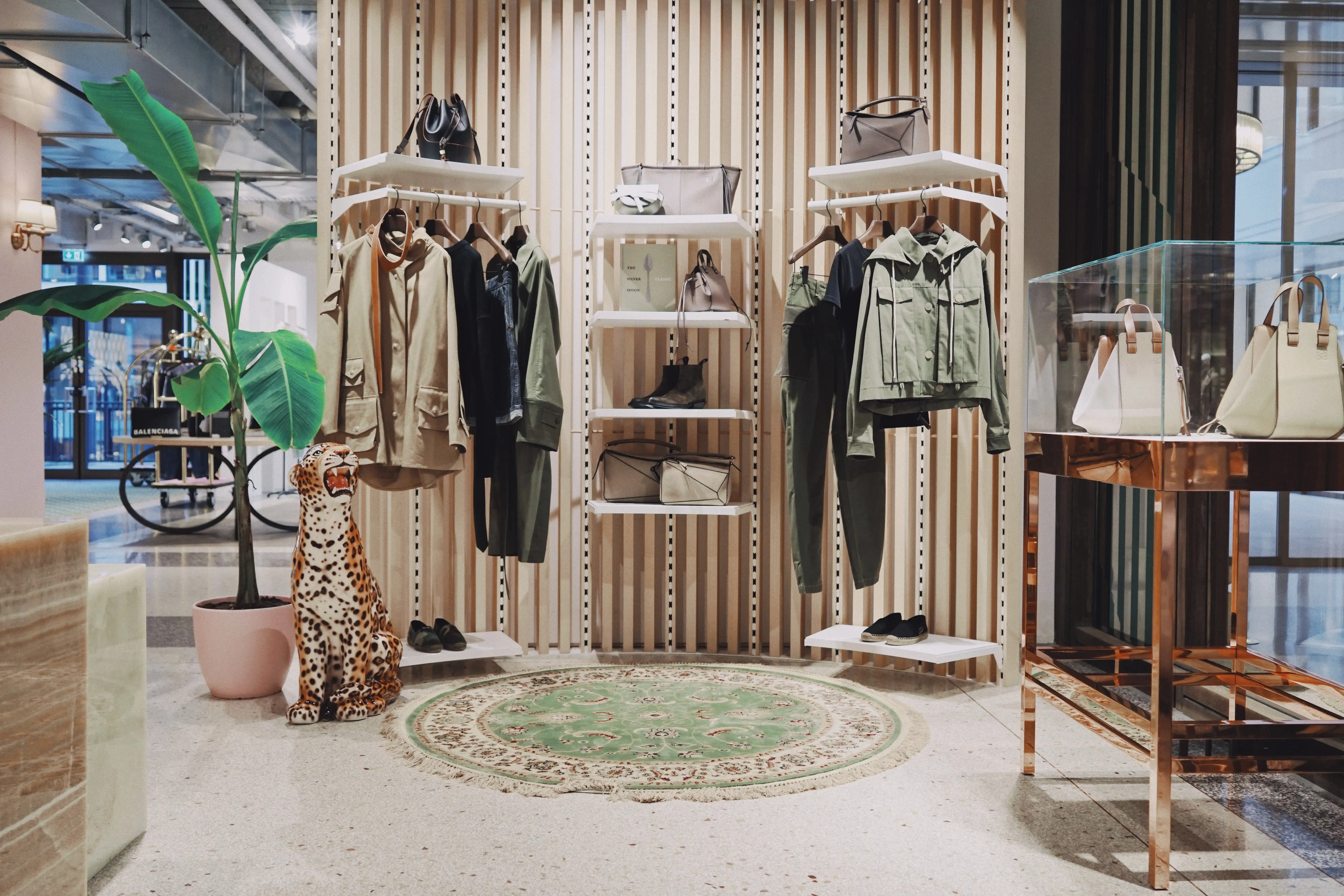 Fashion collection in safari tones at UZWEI FASHION HOTEL on white rails against a wooden slat wall, with a ceramic leopard figure, a round green oriental rug and a copper display case.