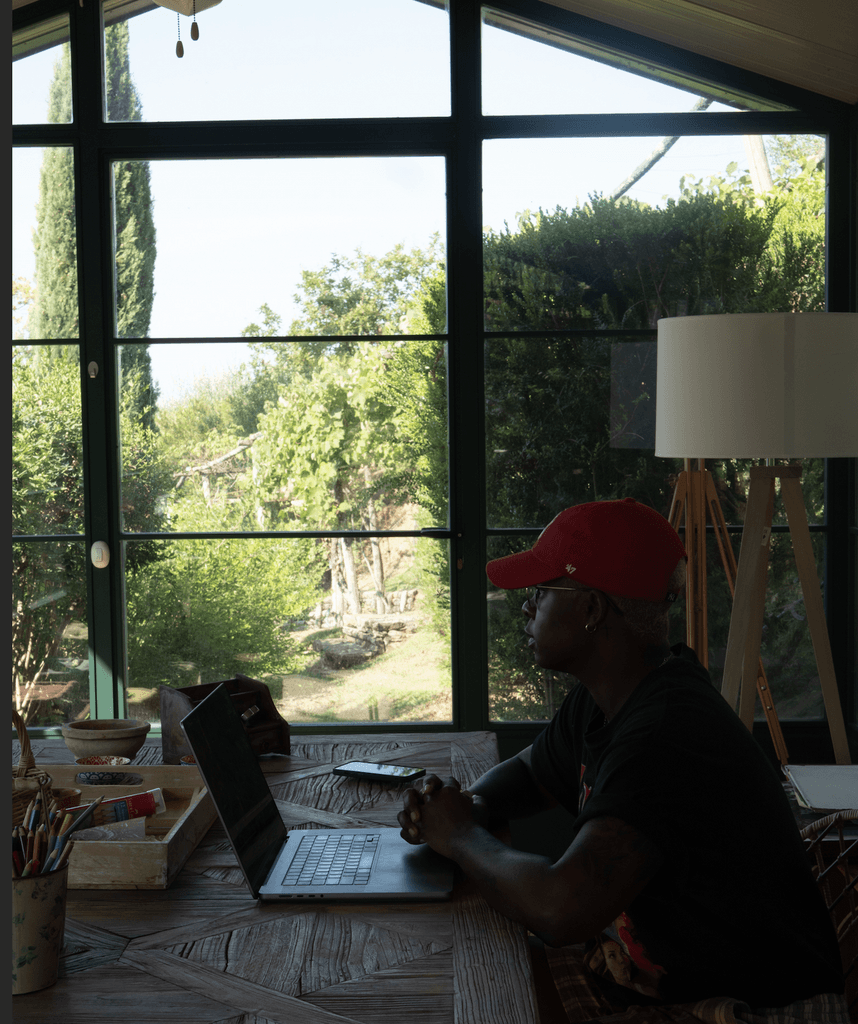 Ashleigh Warren sits at a table working at laptop, looking out through large windows at a green landscape outside.