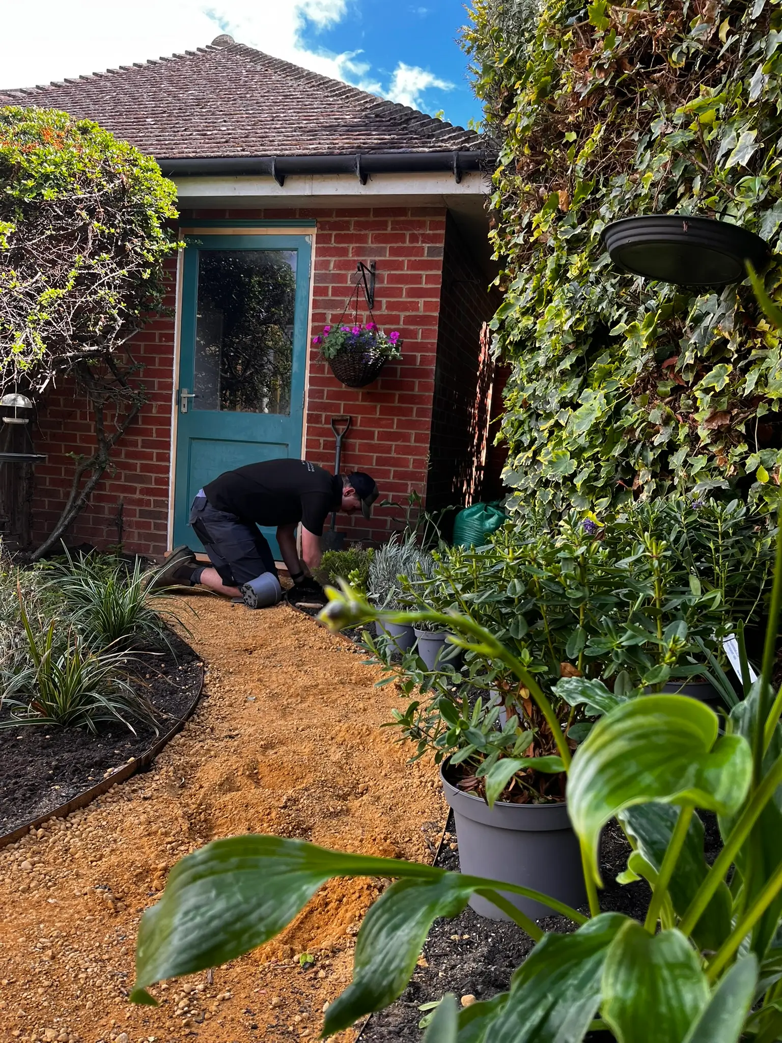 A person gardening near a house surrounded by greenery and a well-maintained pathway.