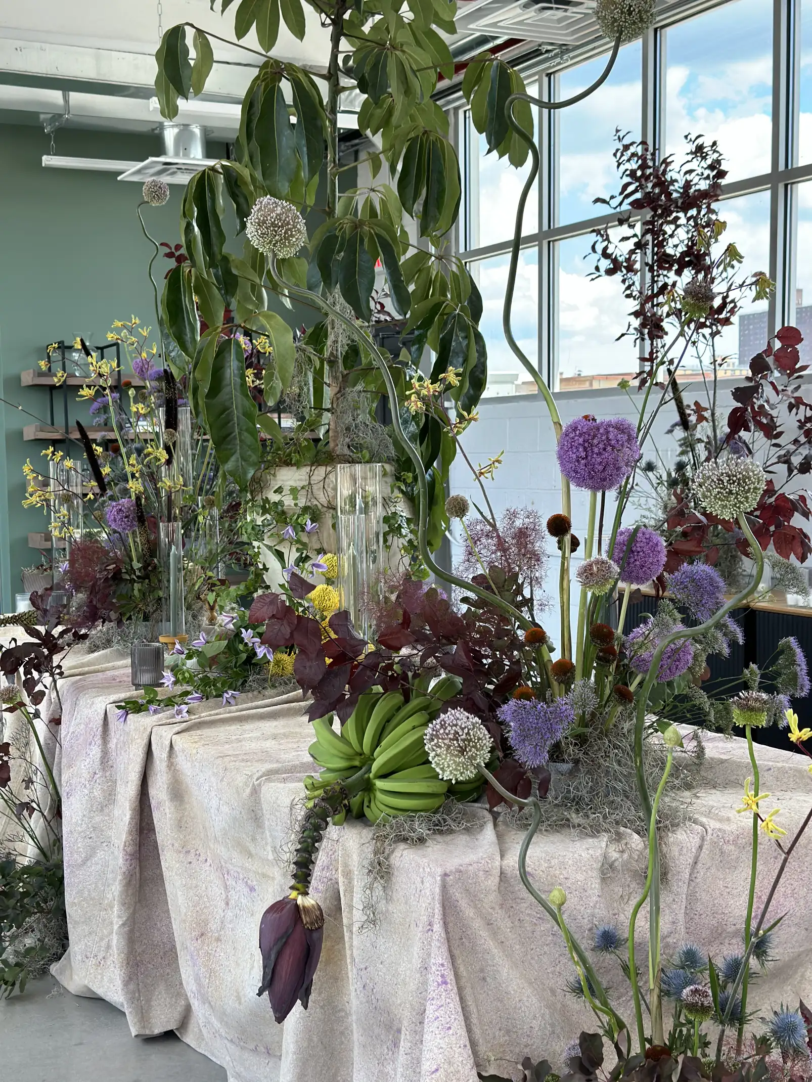 Montaje floral en mesa cubierta con mantel claro, con racimos de bananas verdes, flores moradas y amarillas, follaje rojizo y ramas altas bajo una gran ventana luminosa.
