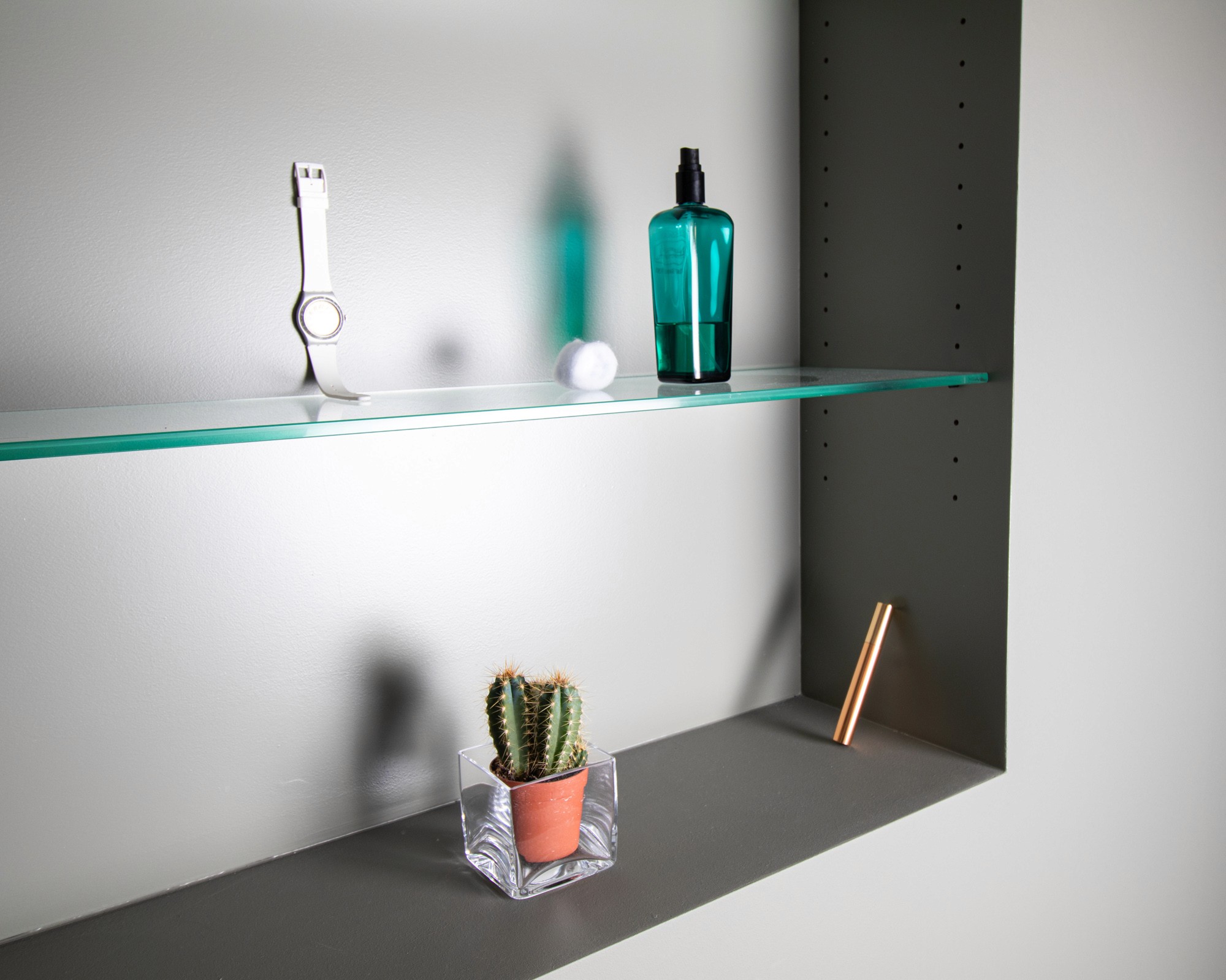 Photographie d'un renfoncement de mur d'une salle de bain comportant une étagére en verre et rempli avec des petits objets tels qu'une montre Swatch, une boutelle de parfum Hermè, un rouge à lèvre et un petit cactus