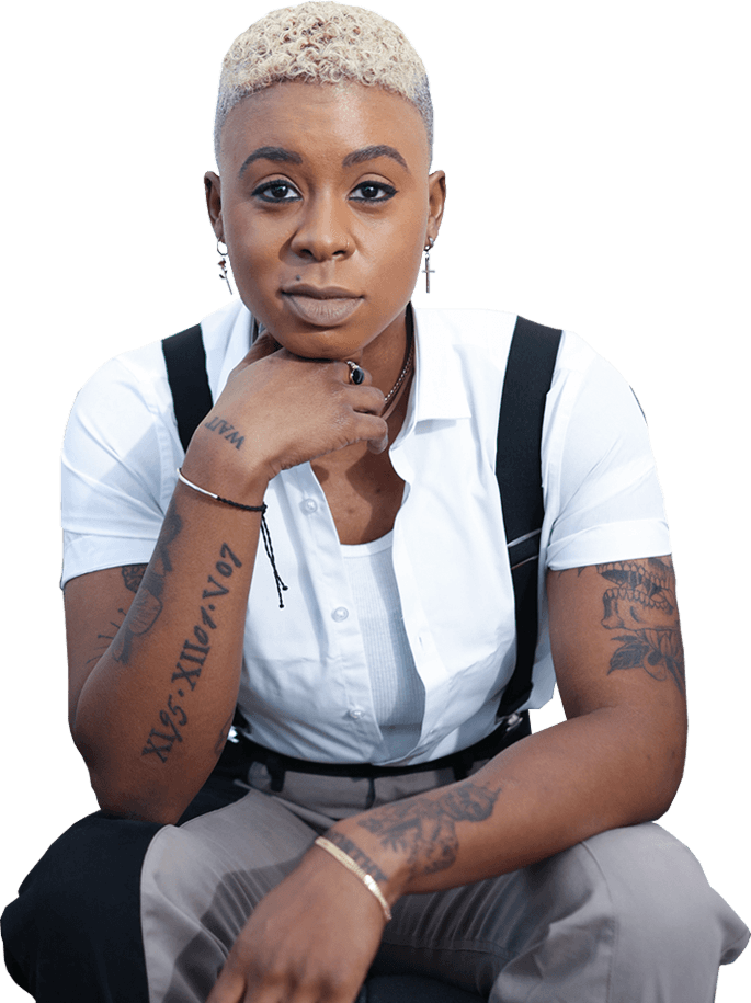 Ashleigh Warren, a person with short blonde hair and tattoos, wearing a white shirt and black suspenders, sitting thoughtfully.
