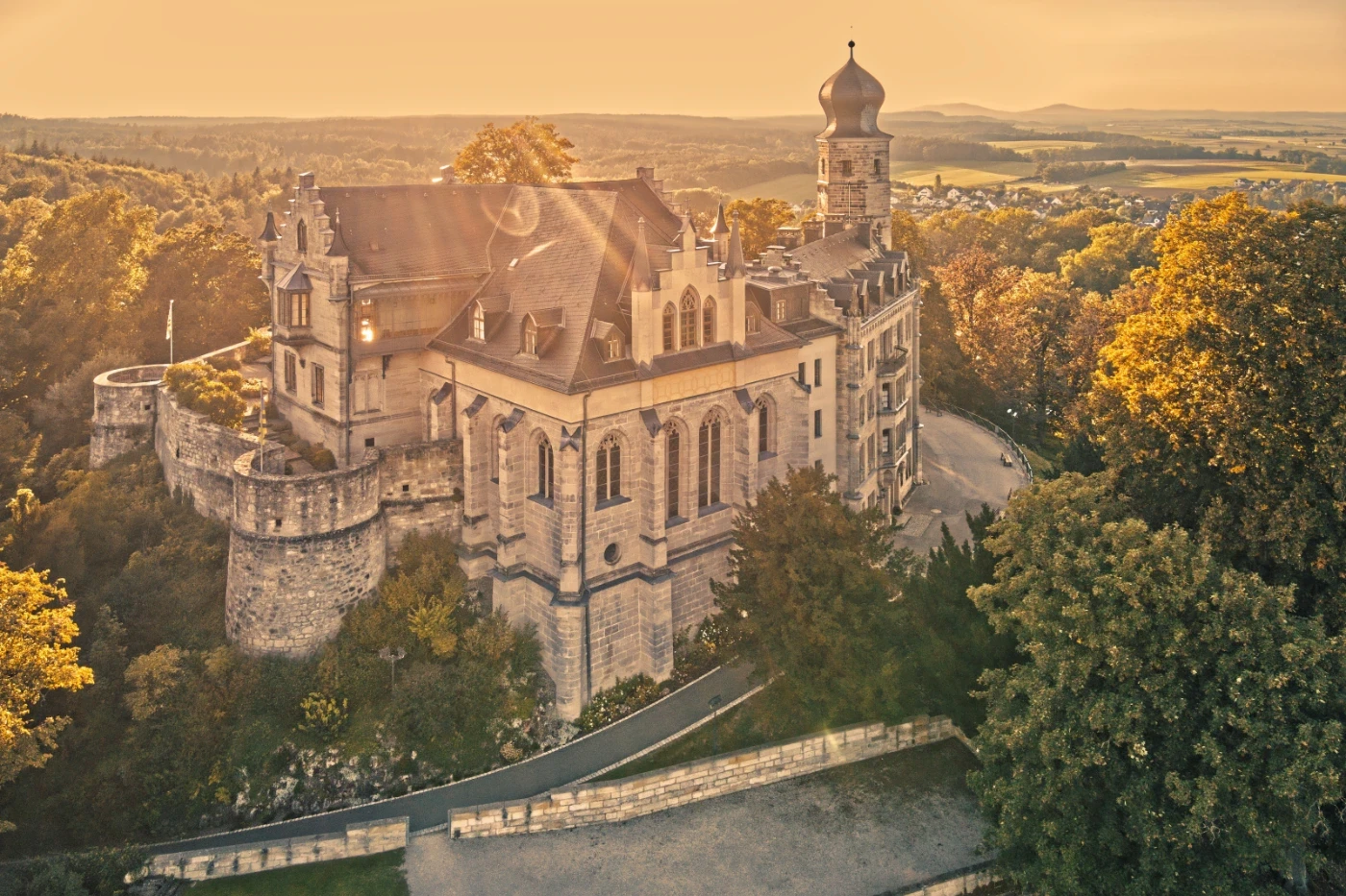 Drohnenaufnahme eines historischen Schlosses mit Türmen und Rundmauer auf einem bewaldeten Hügel bei Sonnenuntergang. Das warme Licht betont Architektur und Umgebung in einer weiten, ländlichen Landschaft.