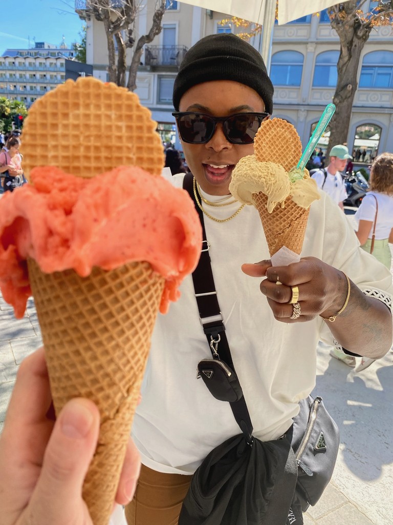 A person holds a cone of orange gelato while Ashleigh Warren smiles and points, also holding a gelato cone.