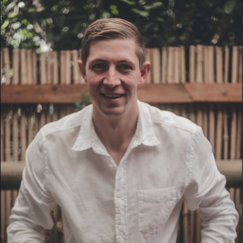 A smiling man with curly hair is sitting at a desk, looking at the camera, with a laptop and a lamp in the background.