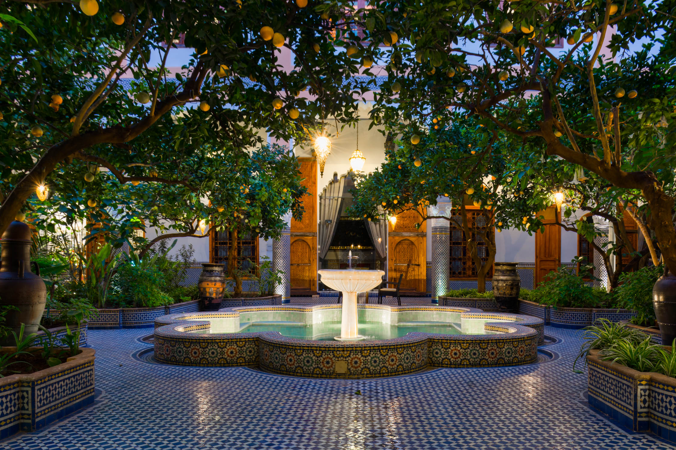 A courtyard with a central fountain and blue tiled floor, framed by orange trees and arches under an evening sky.