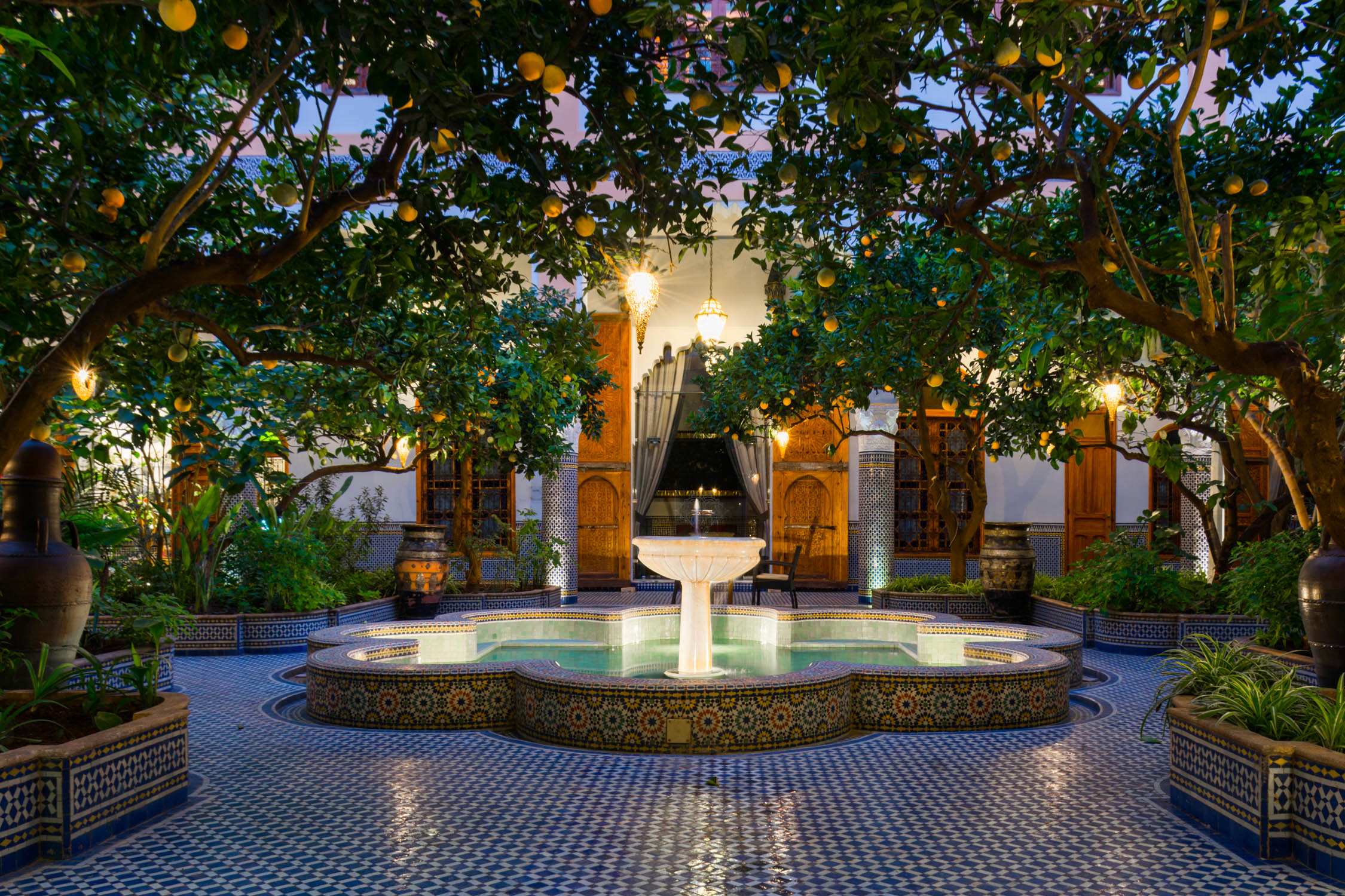 A courtyard with a central fountain and blue tiled floor, framed by orange trees and arches under an evening sky.