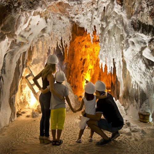A family wearing hard hats explores a cave with hanging stalactites and an illuminated orange rock formation in the background.