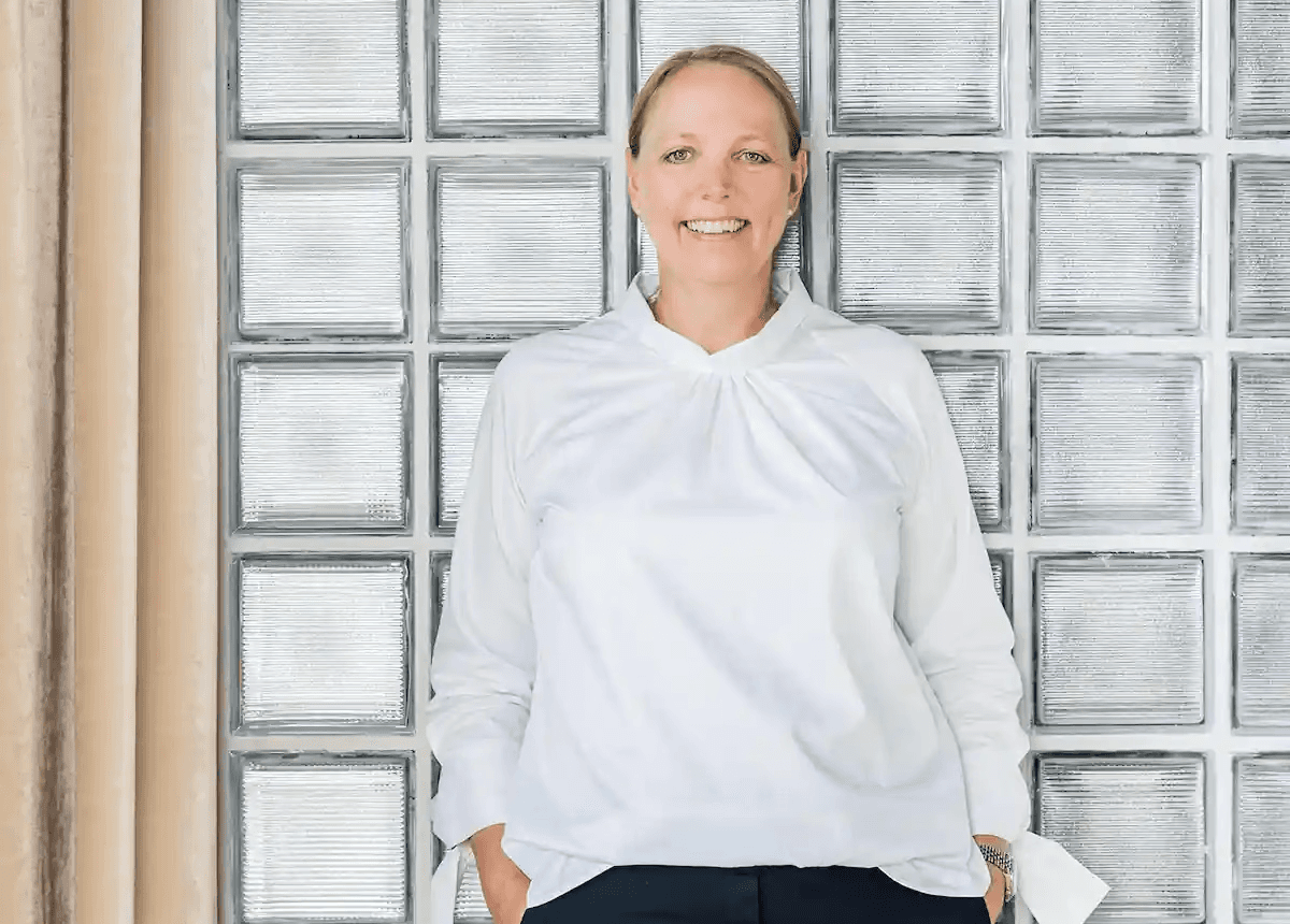 Person mit zusammengebundenen Haaren trägt eine weiße Bluse und dunkle Hose, steht im Büro vor einer Glasbausteinwand und lächelt