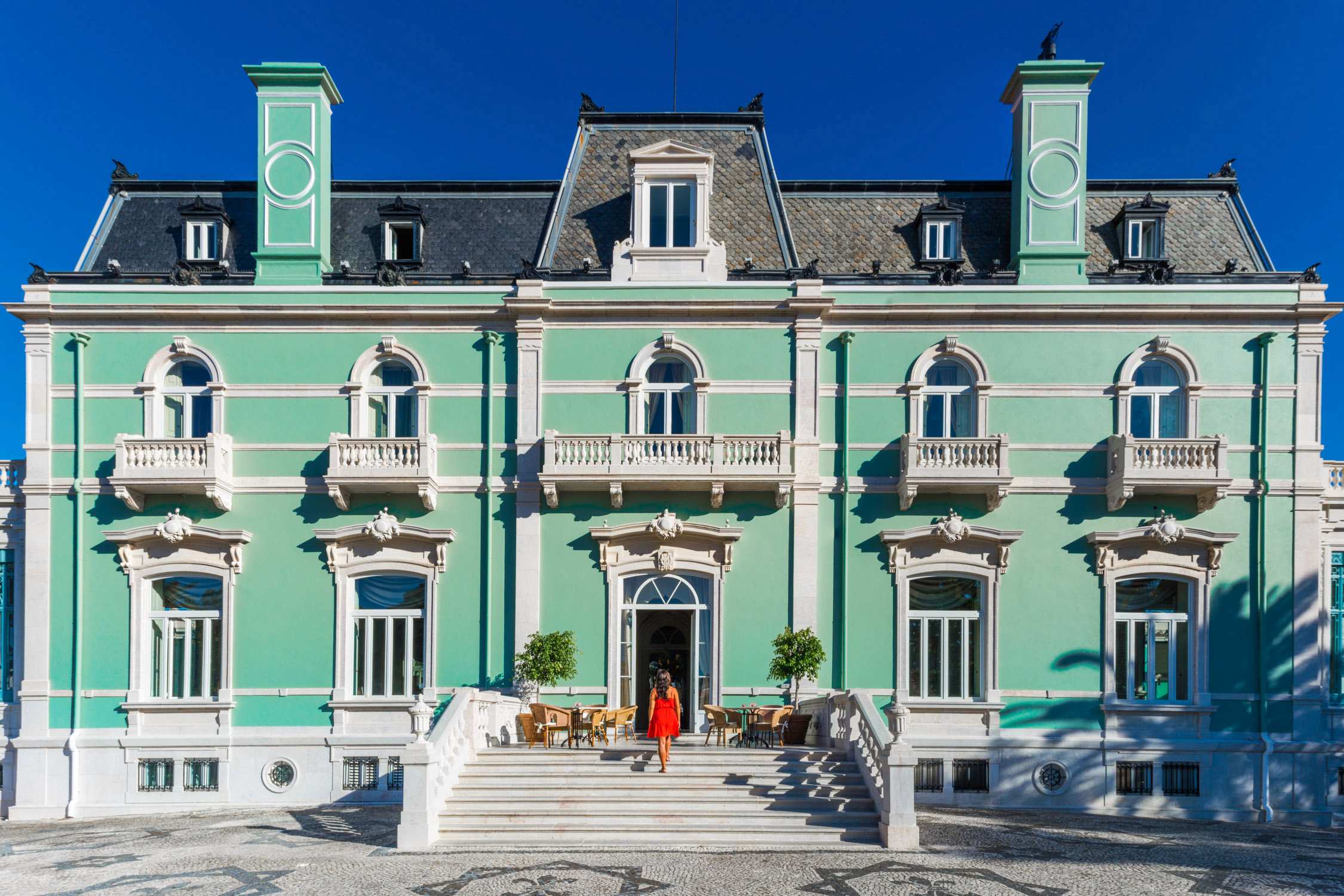 A woman in a red dress stands on the steps of a mint green and white French-style mansion under a clear blue sky.