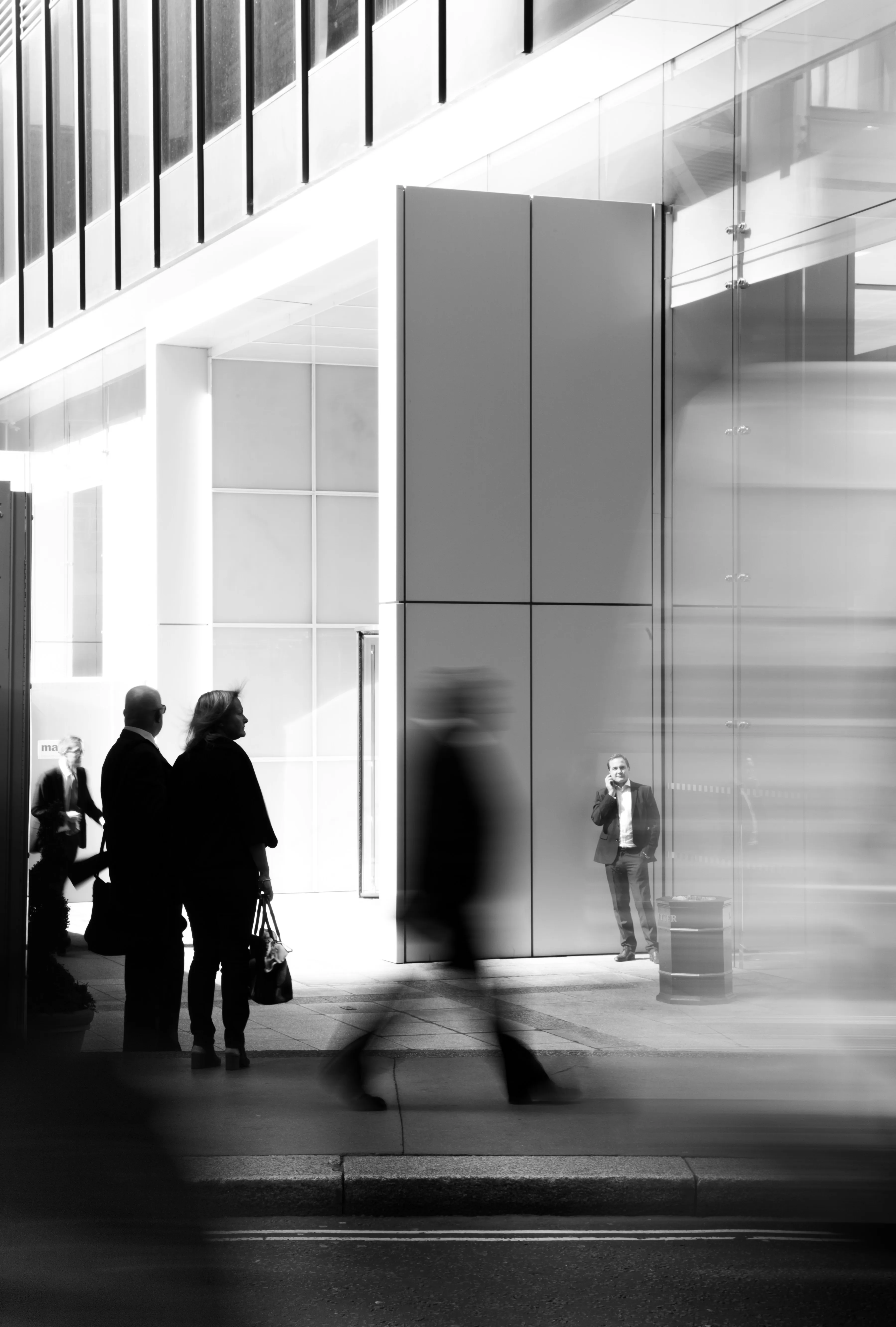 Silhouetted figures walk past a modern building in a blurred, monochrome cityscape.
