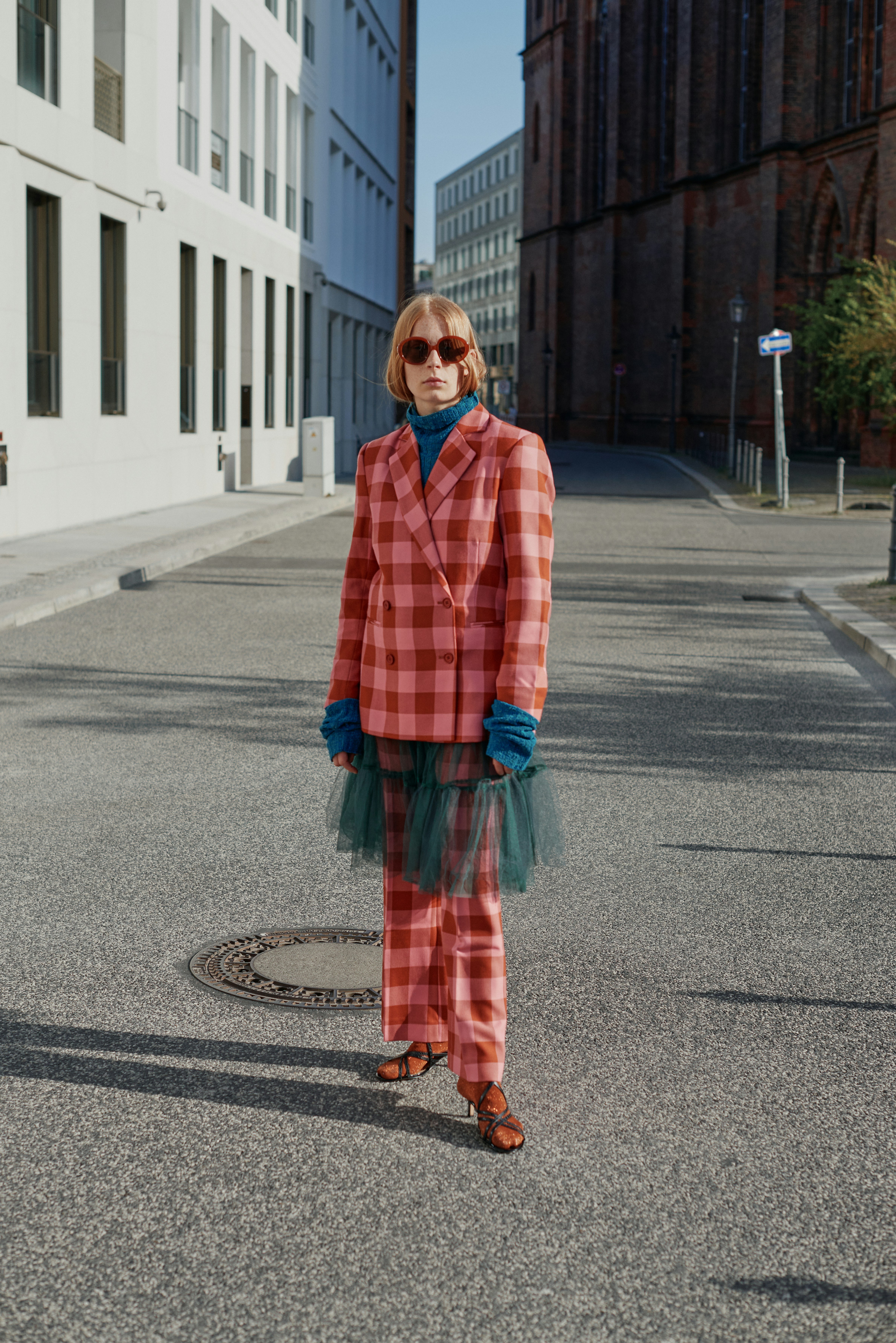 Full-length editorial street portrait of a blonde model in a pink and red checked suit with teal tulle hem, standing on an empty urban street