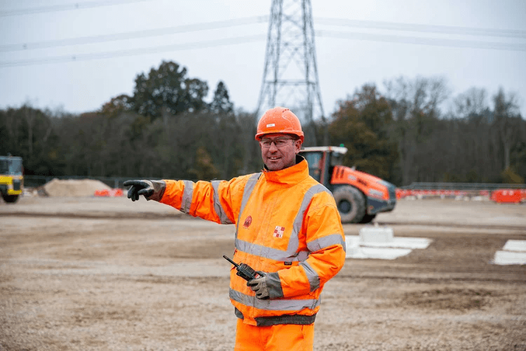 Person in orange jacket pointing at something on a tripod.