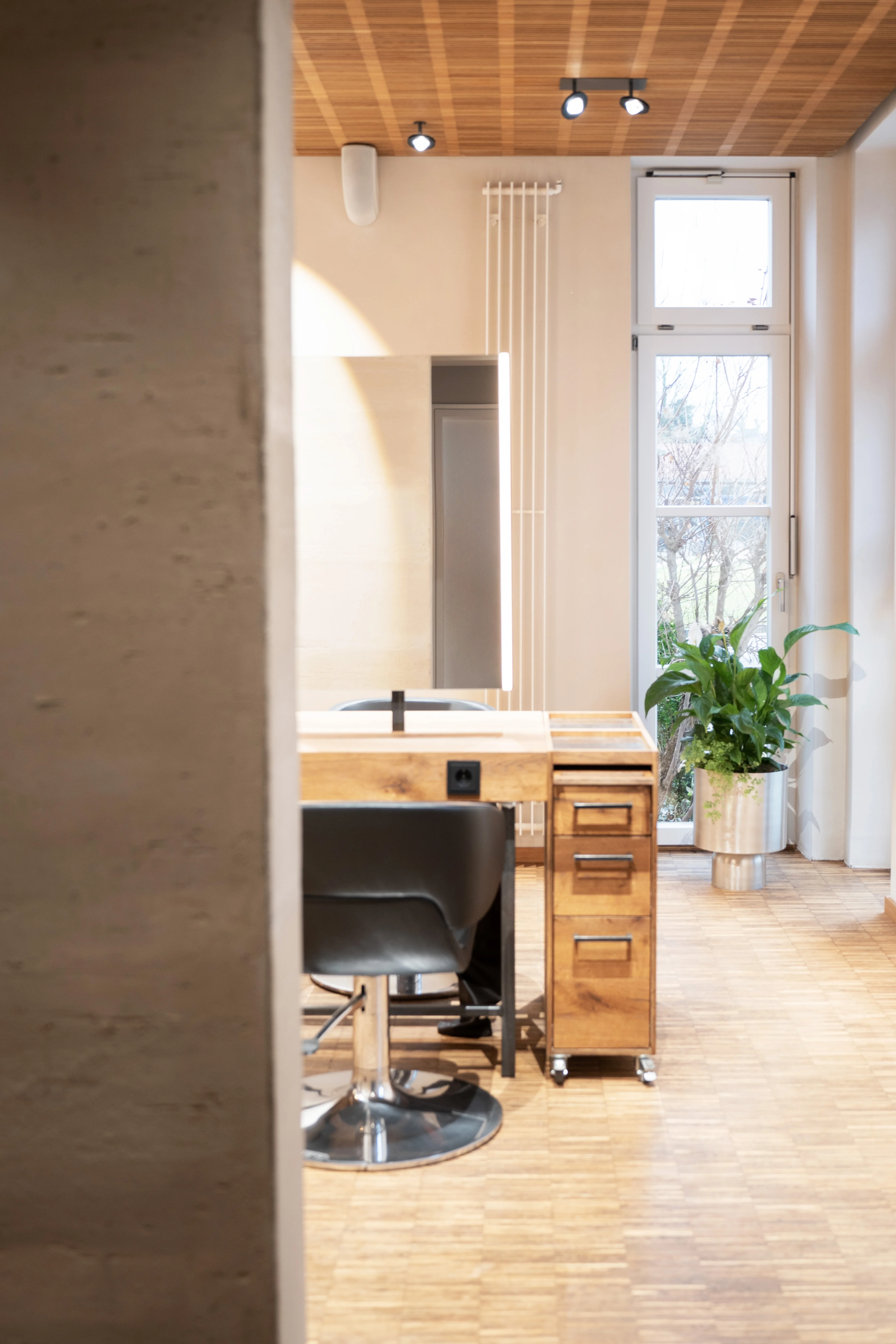 Single styling station at EMPL FRISEURE with a black salon chair, oak counter with mirror and a vintage wooden drawer cart, next to a tall window with plants.