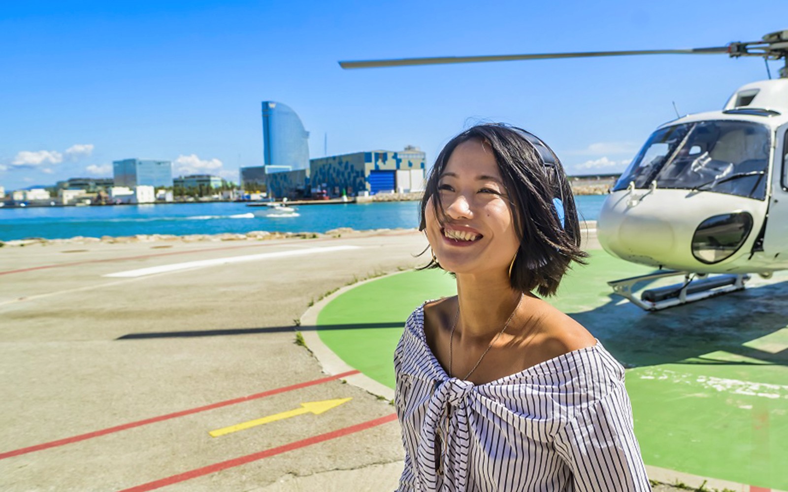 一位微笑的女士站在直升机旁，背景是巴塞罗那的天际线。