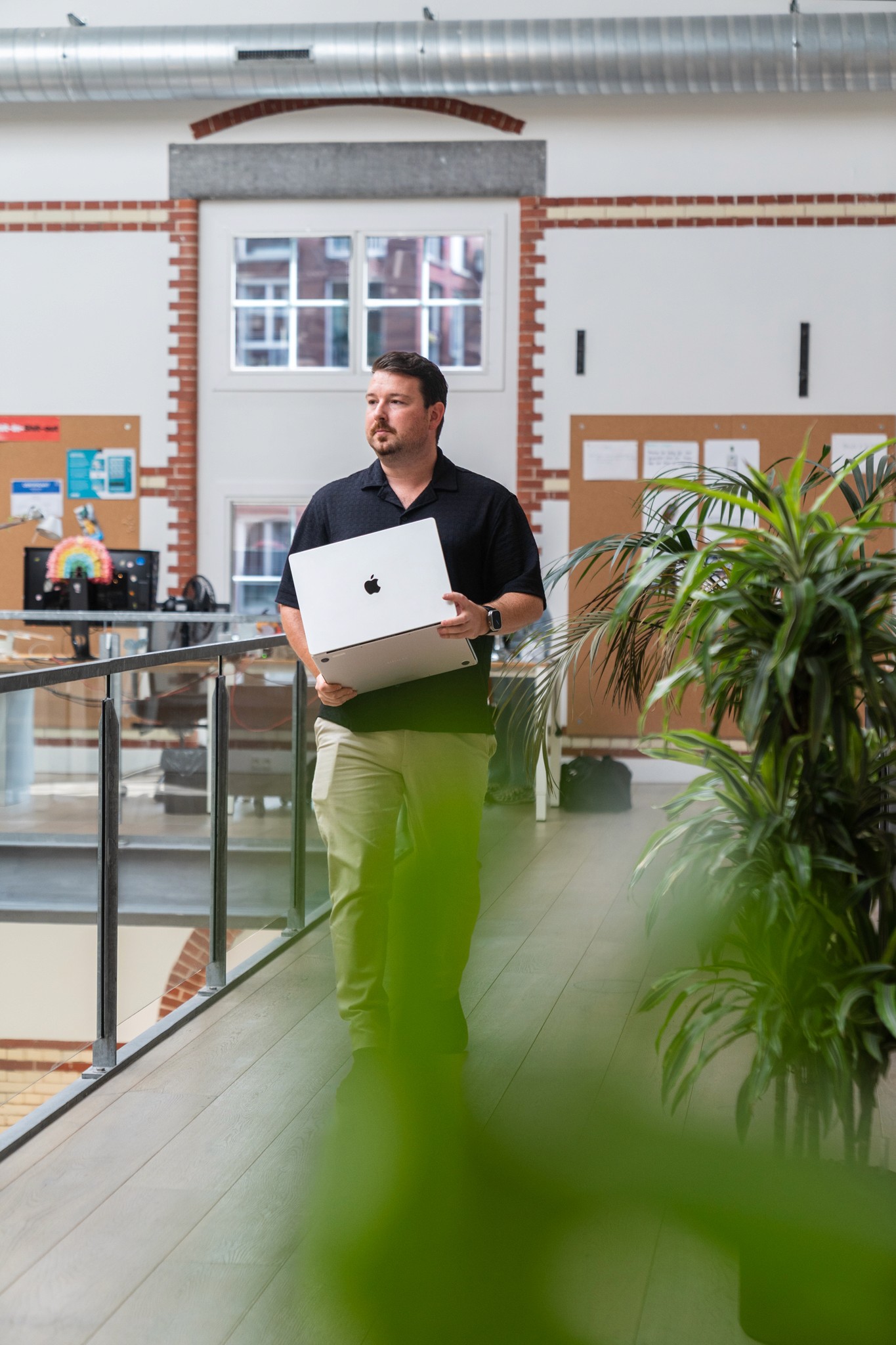 A man holding a laptop, walking past plants in an office environment