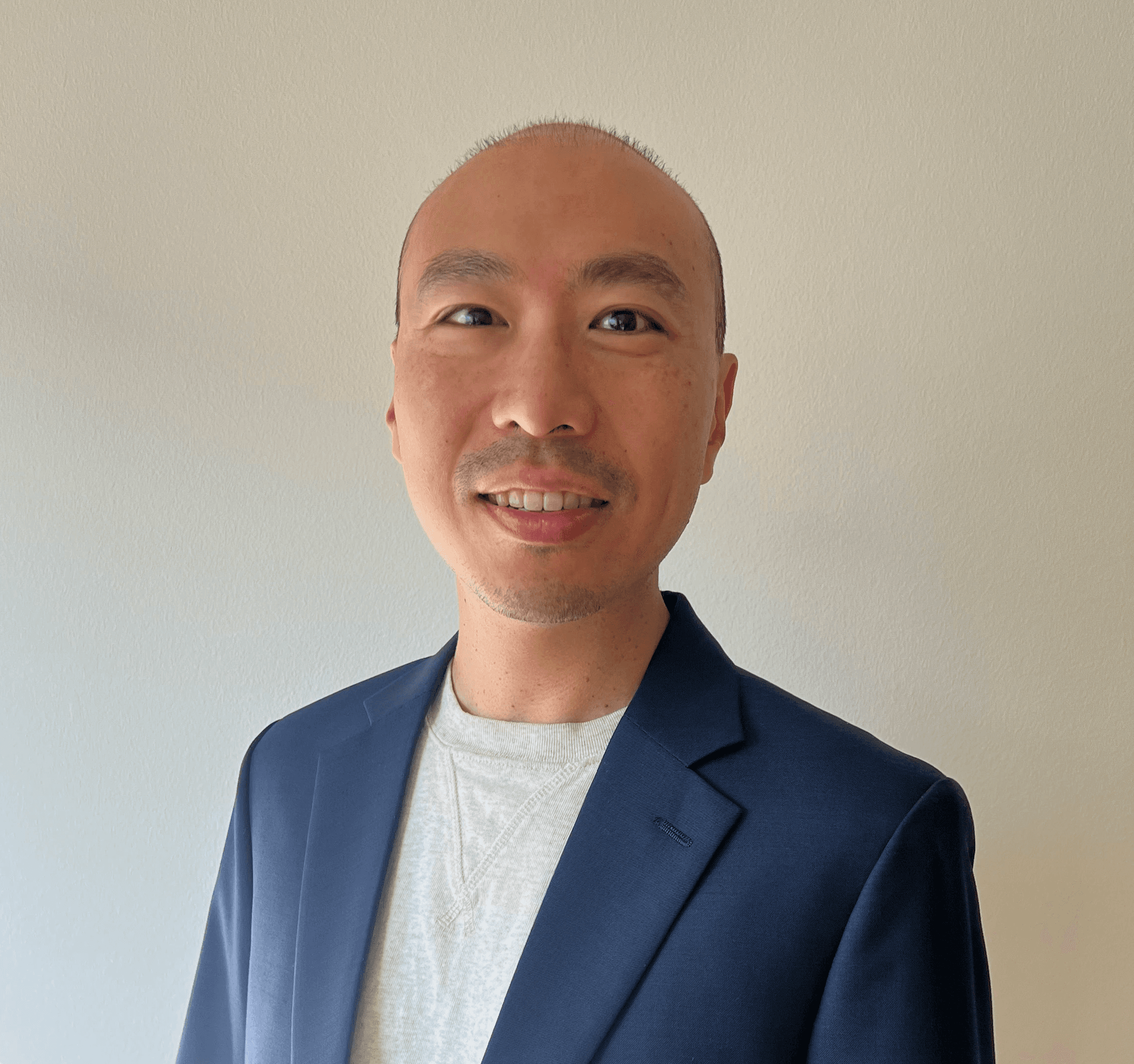 A smiling person with curly hair sits at a desk, looking at a laptop against a dark background.