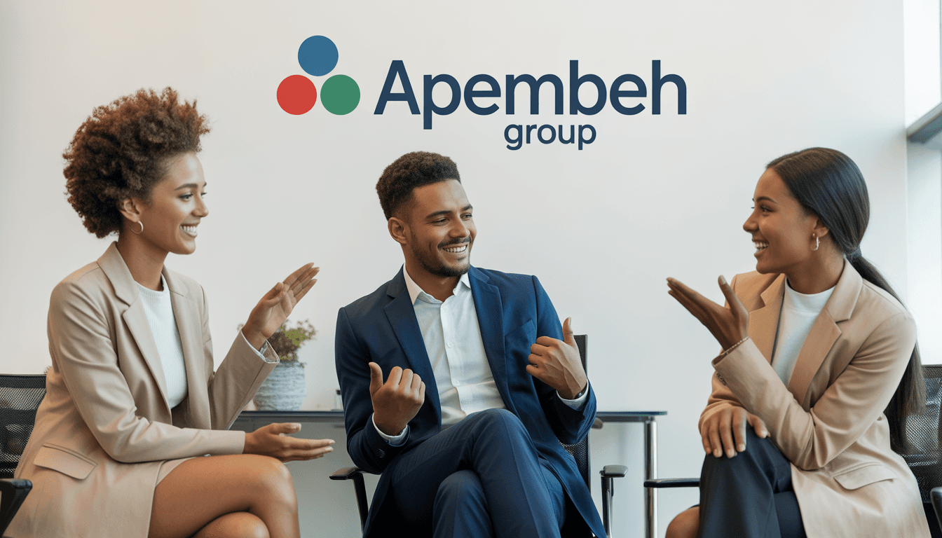 Two people sit at a white table in a dimly lit room, discussing something. The man gestures towards a laptop, while the woman listens attentively.