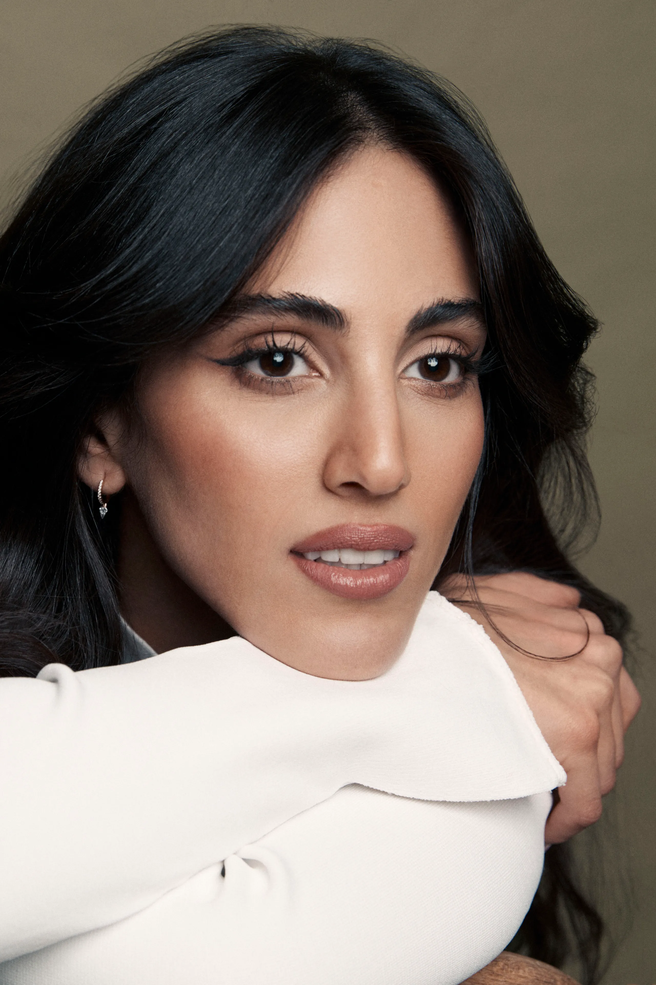 Close-up portrait of a young woman with green eyes, natural makeup, and dark hair, gently touching her face, against a neutral beige background