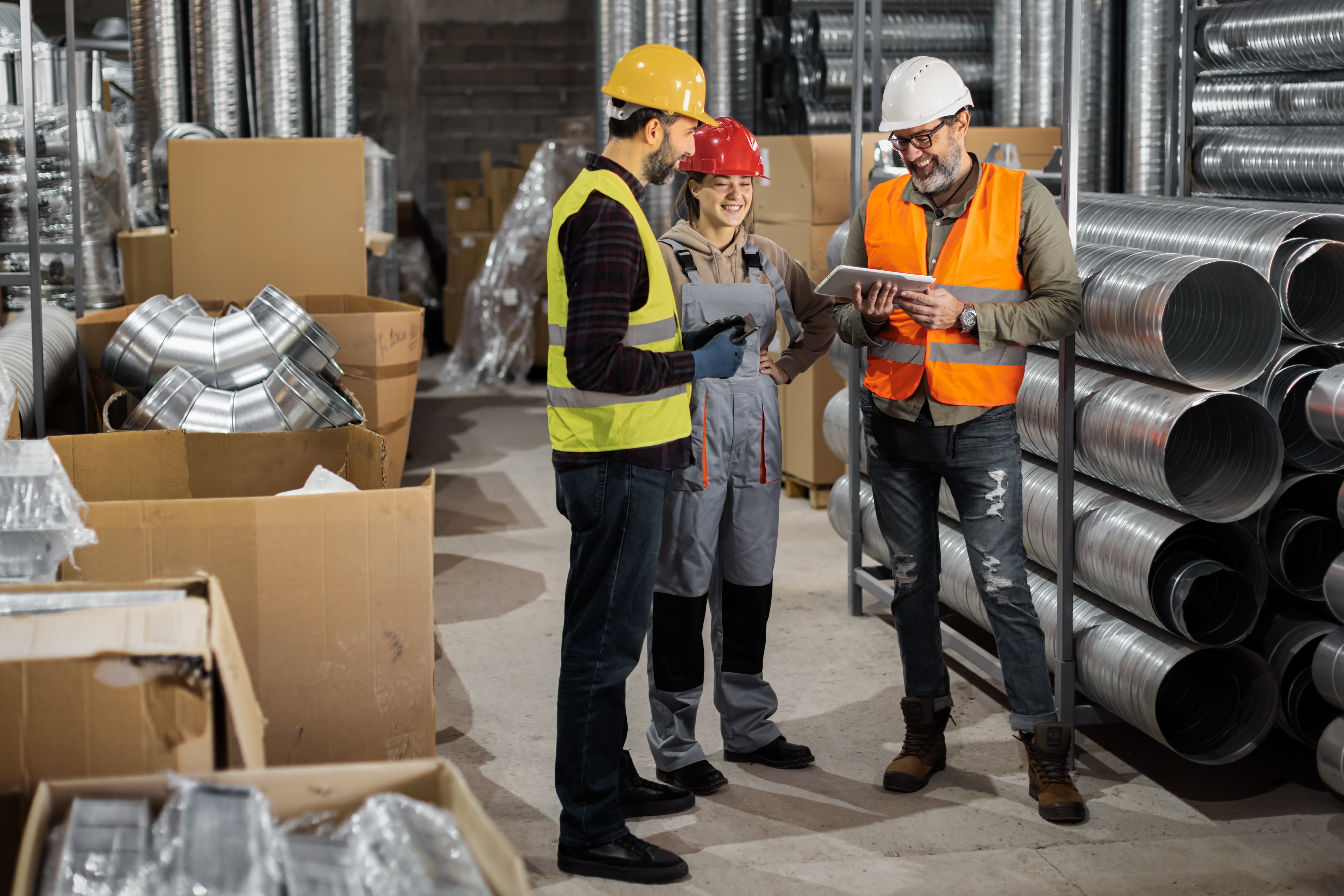 Three construction workers in safety gear examine plans in a warehouse filled with materials and equipment.