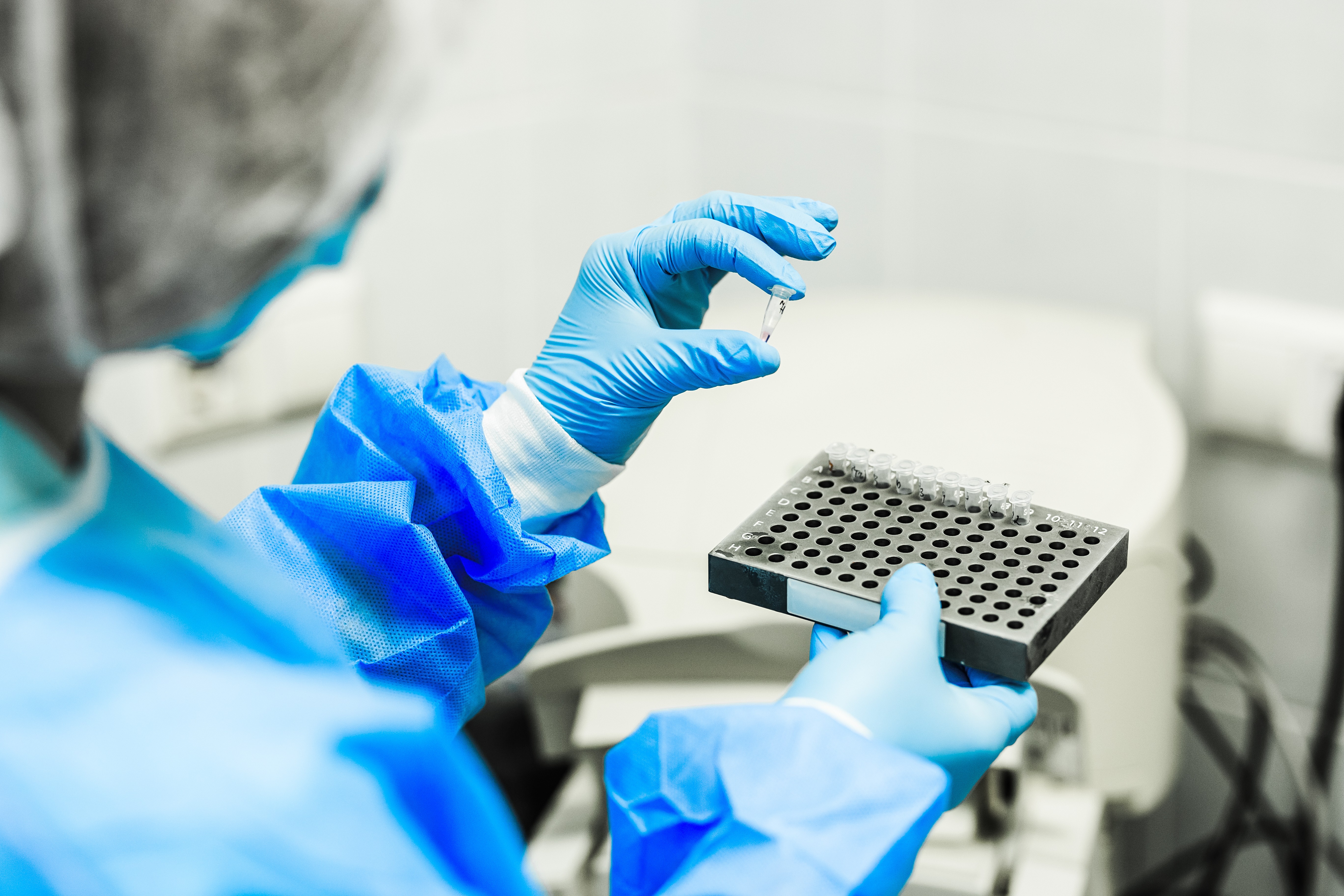 A scientist holding a vial in their left hand, and a tray of vials in their right hand. 