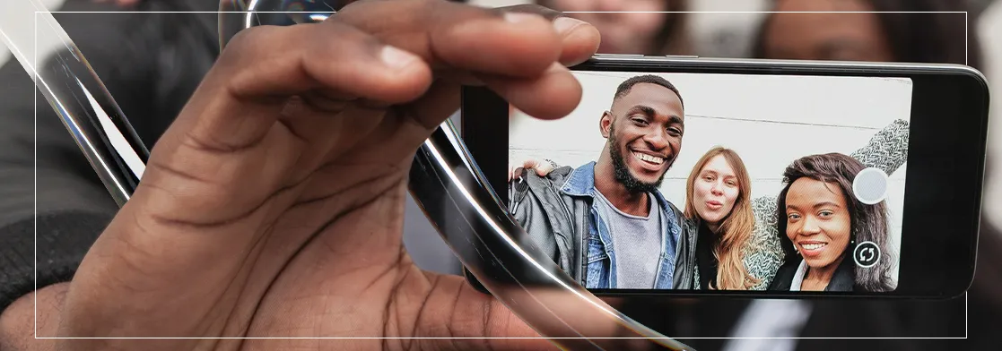 Close de uma mão segurando um smartphone que exibe três amigos sorridentes tirando uma selfie.