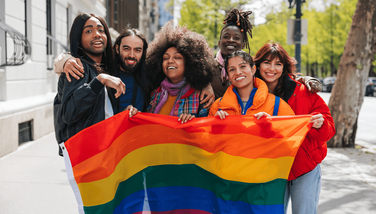 A group of people holding the pride rainbow flag standing outside on the sidewalk 