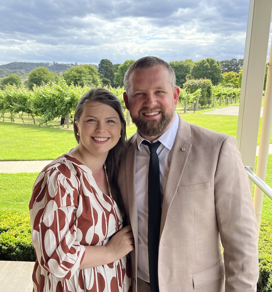 A smiling couple poses together outdoors in front of a lush vineyard, with overcast skies above and vibrant greenery in the background.