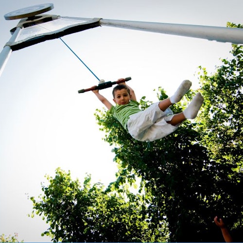 Child in a striped shirt swings high on a zip line, with trees and a clear sky in the background.