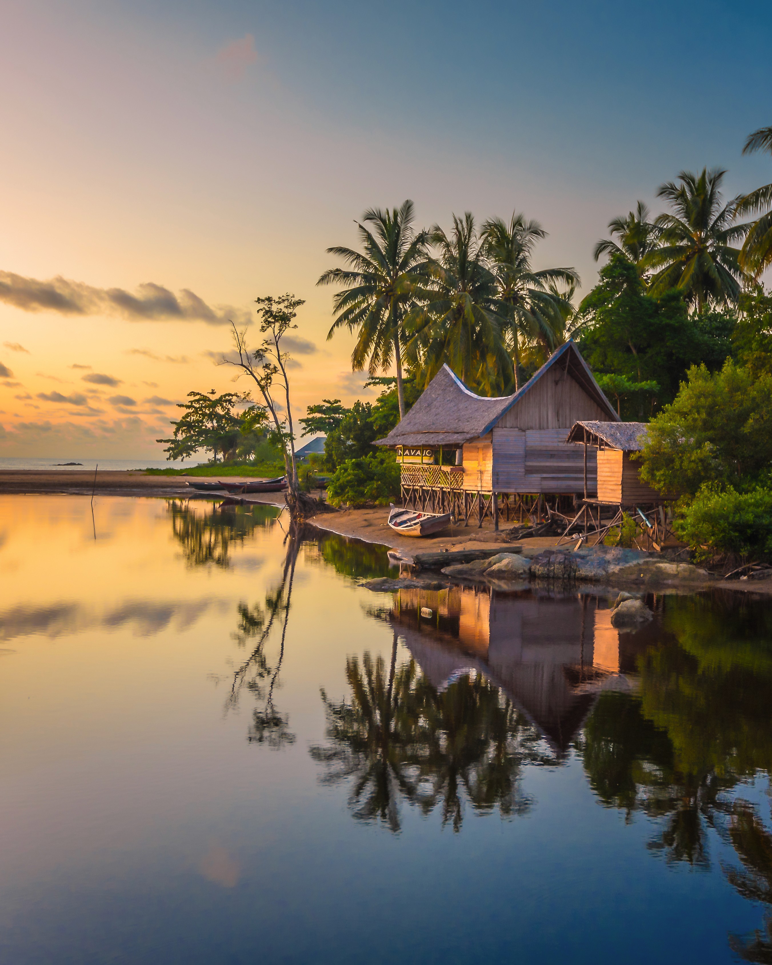 Tropical sunset scene featuring a thatched-roof hut by calm water, reflecting palm trees and a colorful sky. The mood is tranquil and serene.