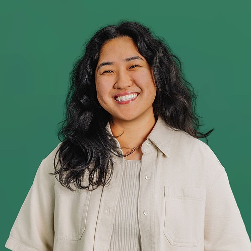 Smiling woman with wavy hair wearing a light-colored button-up shirt, standing against a green background.