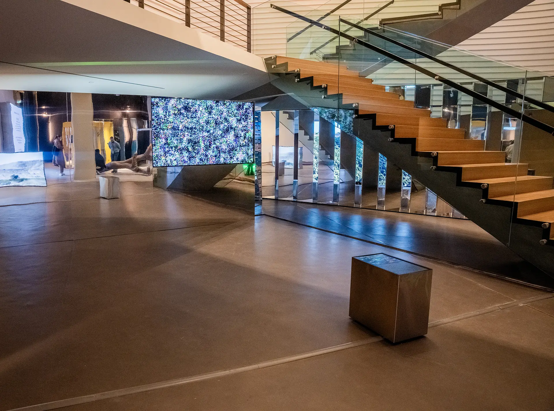 A contemporary art gallery interior featuring a large digital screen displaying colorful static noise, situated next to a modern wooden staircase with glass railings. The minimalist space includes polished concrete floors, mirrored wall panels reflecting digital art, and visitors exploring interactive exhibits in the background.