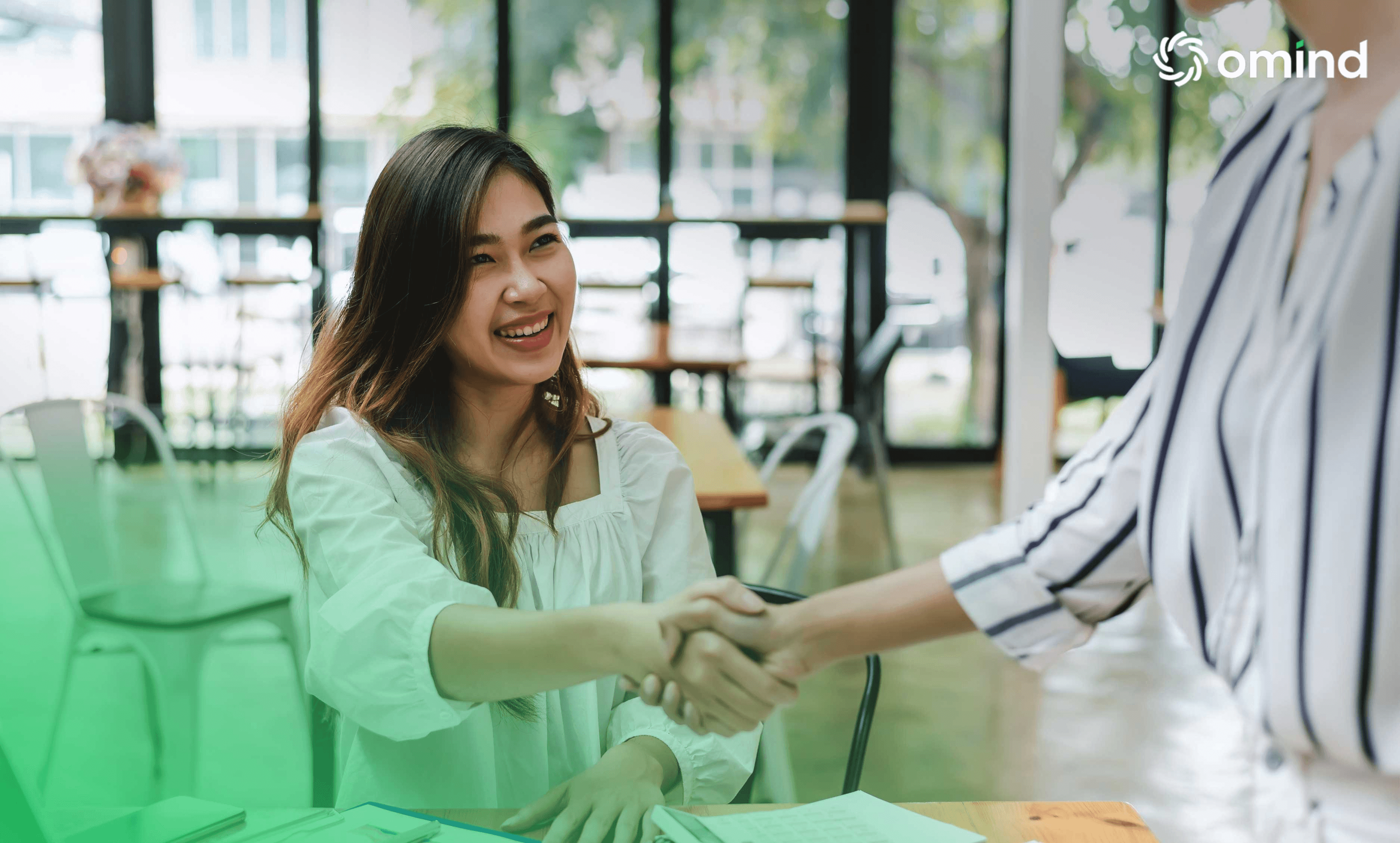 A smiling woman shakes hands with another person in a bright, modern office setting.