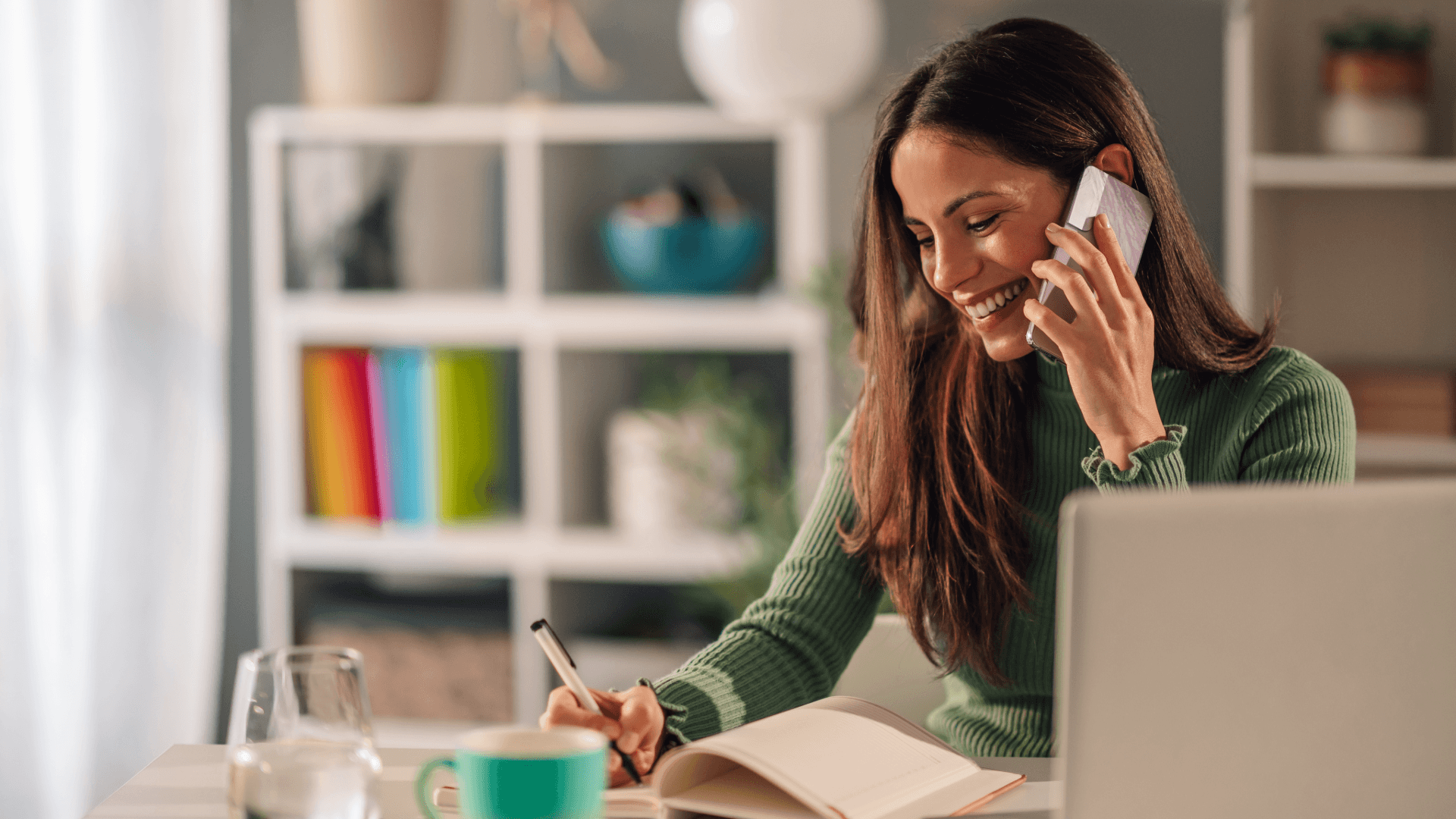 Mujer sonriente hablando por teléfono con un agente de e-commerce, en su escritorio con un ordenador y un cuaderno.