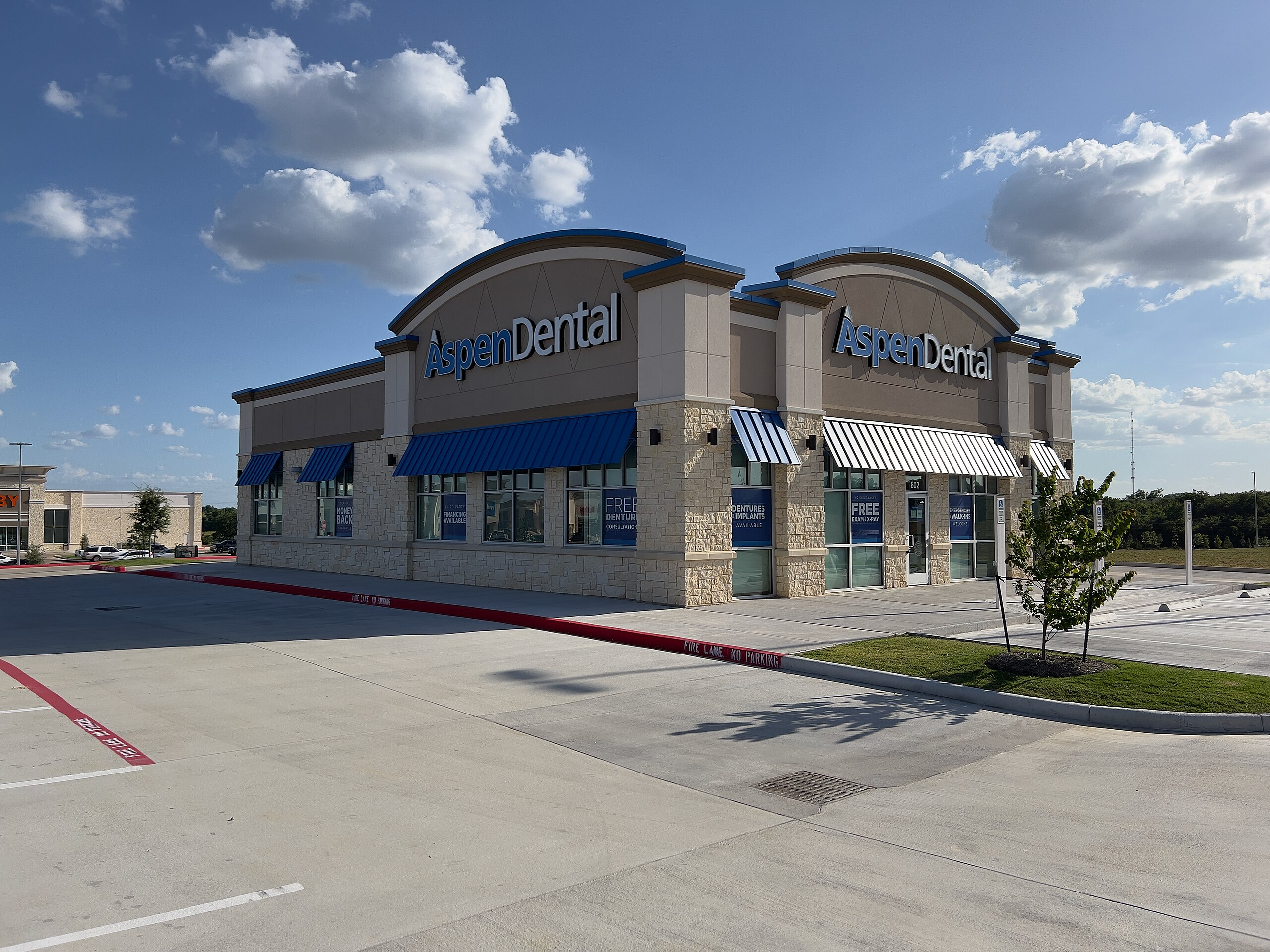 A modern Aspen Dental clinic, built by Meadway Construction, features blue awnings and large windows. The mostly empty parking lot sits under a partly cloudy sky, with window signs offering free exams and X-rays for new patients.